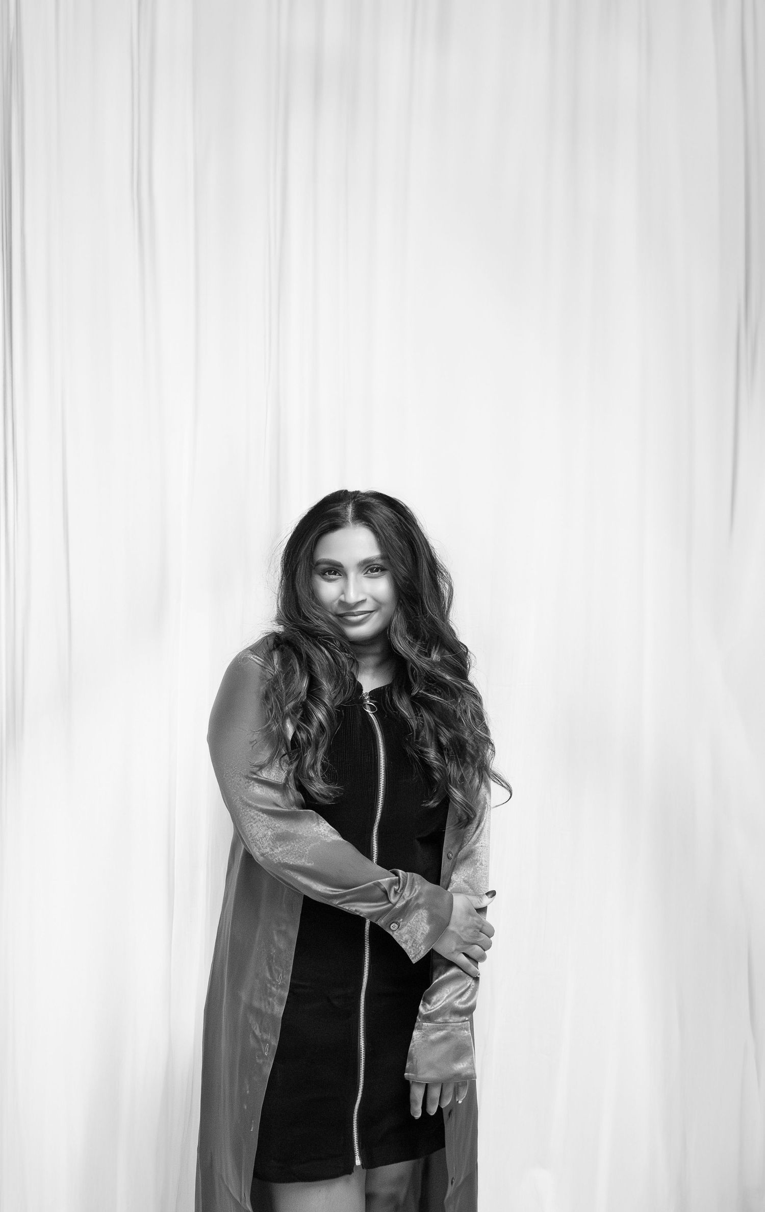 Black and white portrait of someone posing in a long sleeve jacket over a short dress against a light background.