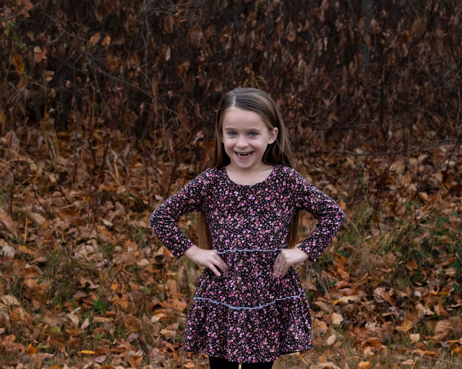 A child in a floral dress poses with hands on hips against a backdrop of autumn leaves and brown foliage.
