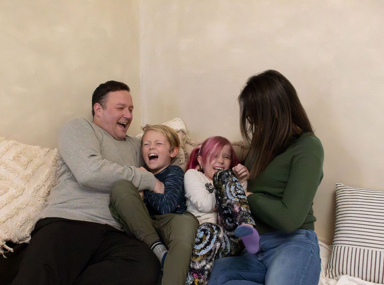A family laughs together while sitting on a couch with decorative throw pillows in a casual home setting.
