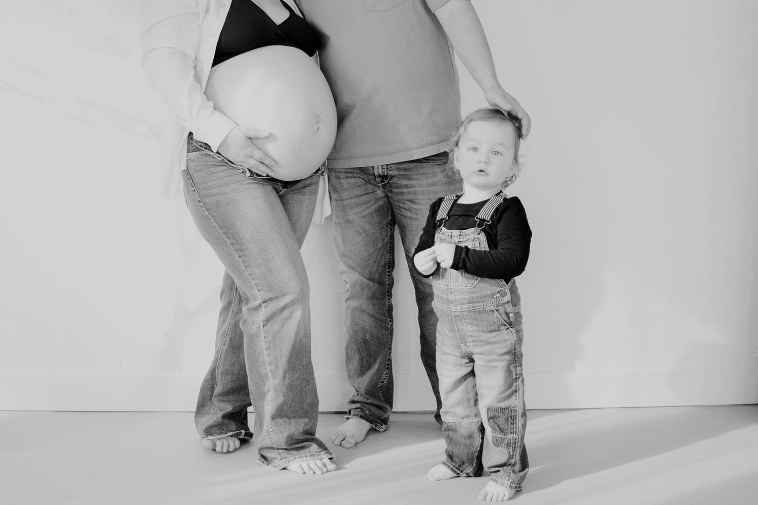 Black and white maternity photo showing an older toddler standing next to pregnant family member, hands on belly.
