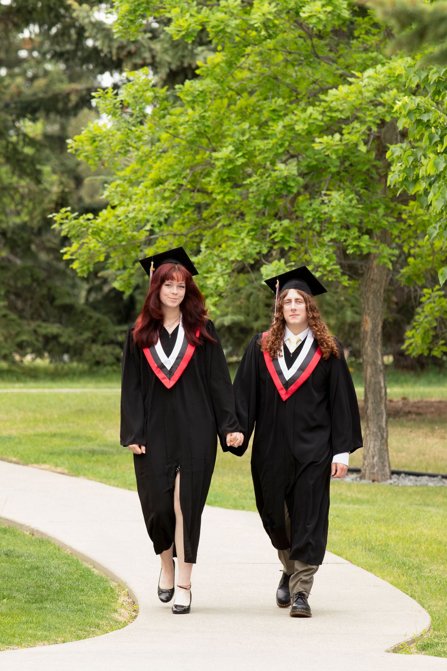 Two graduates in black academic regalia holding hands while walking down a path surrounded by green trees.