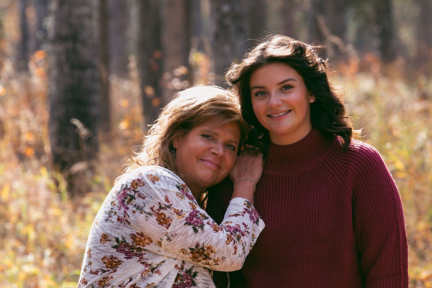Mother and daughter smile together outdoors in autumn with warm sunlight and fall foliage in the background.