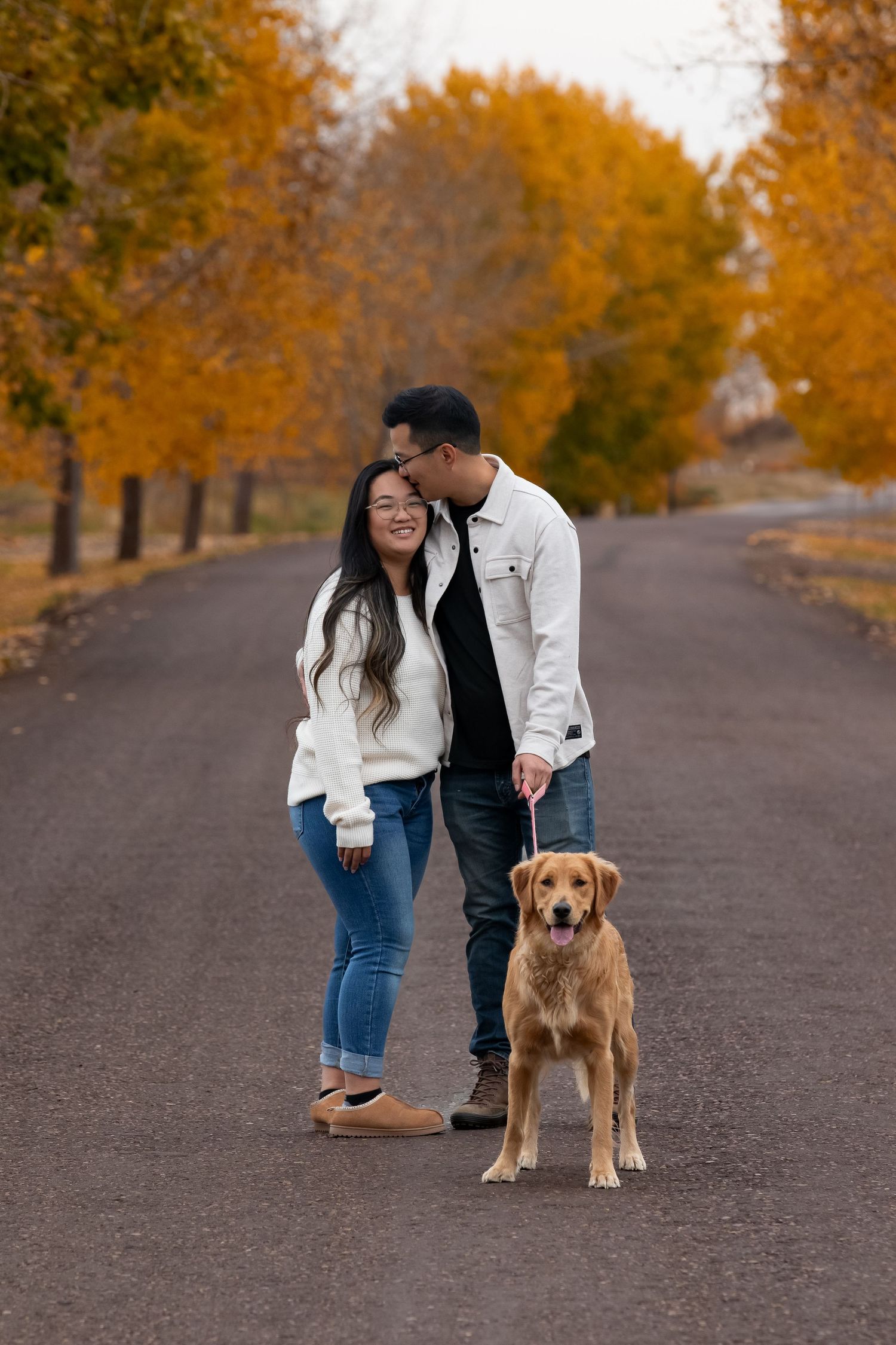 A couple in white sweaters taking their golden retriever dog for a walk on a tree-lined path during autumn.