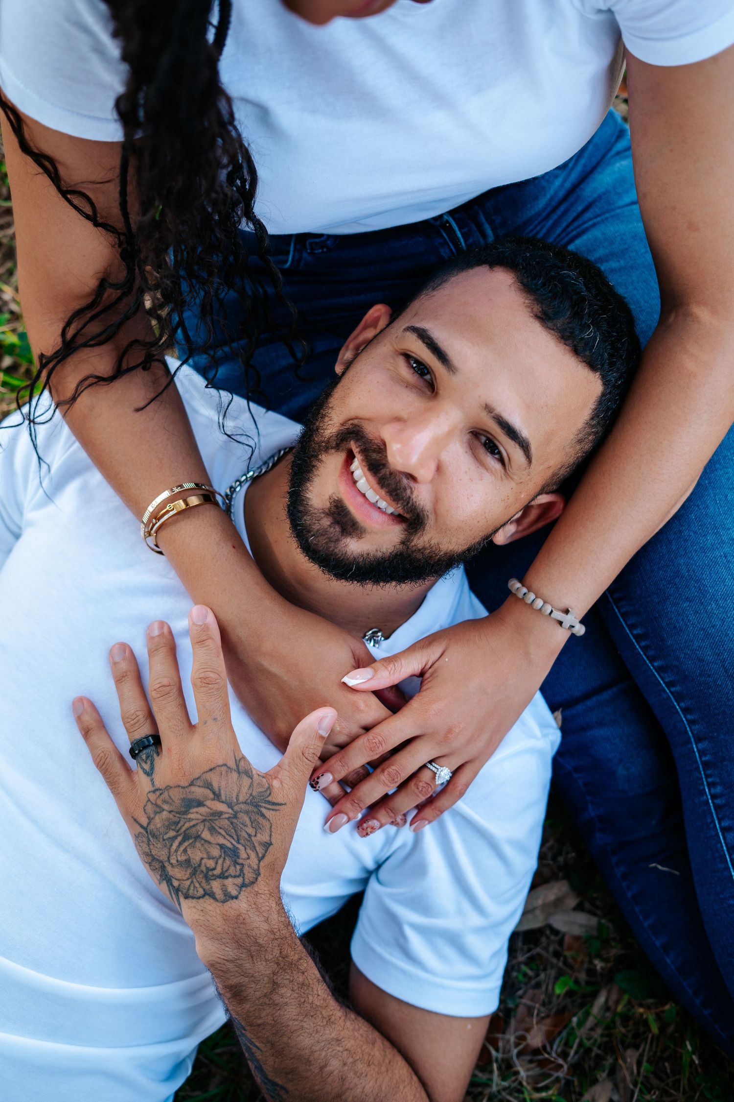 Smiling bearded man lying on a woman's lap outdoors, both wearing white shirts and jeans, showcasing tattoos and jewelry.