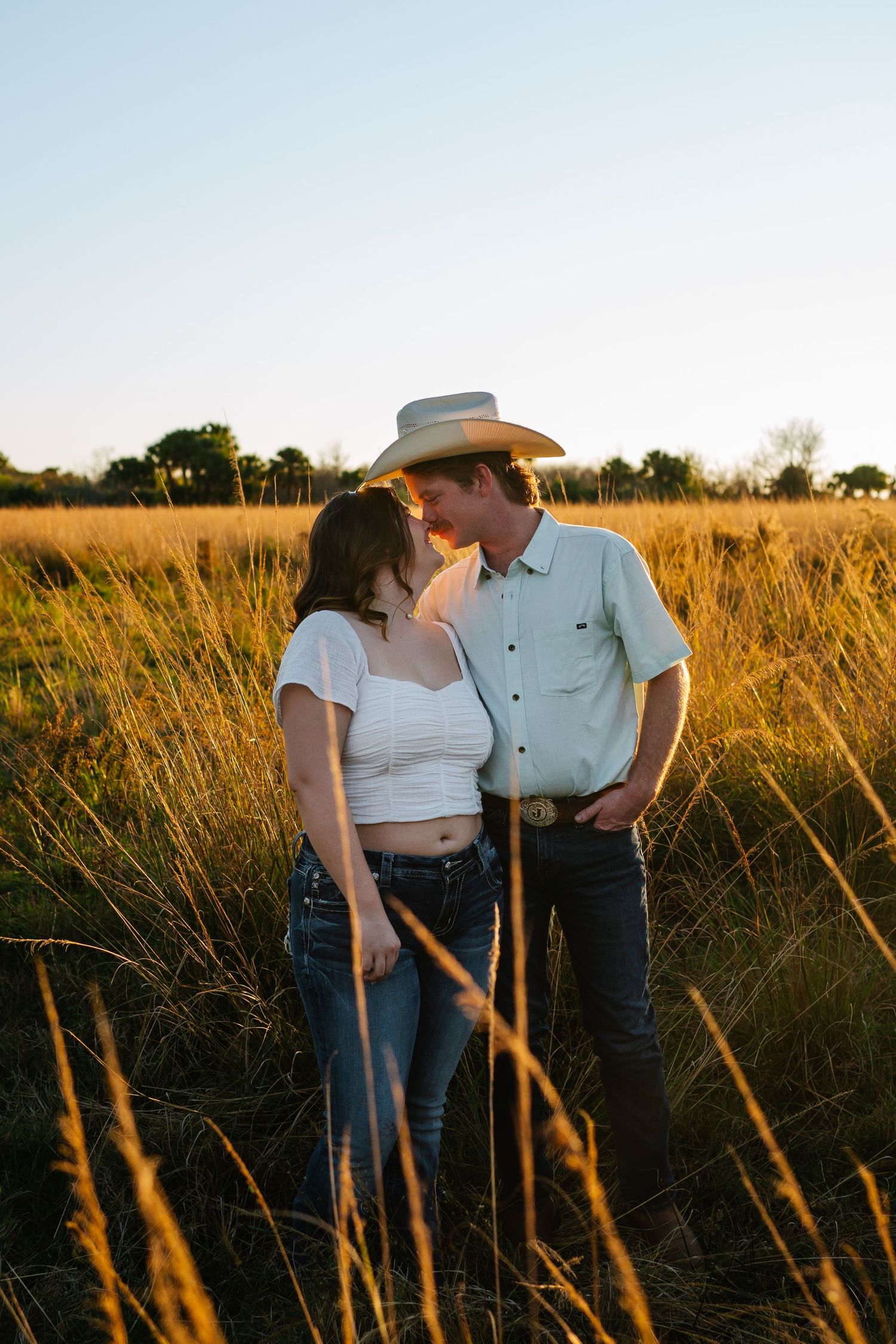 Couple sharing a romantic moment in golden field at sunset, man wearing cowboy hat and light blue shirt.