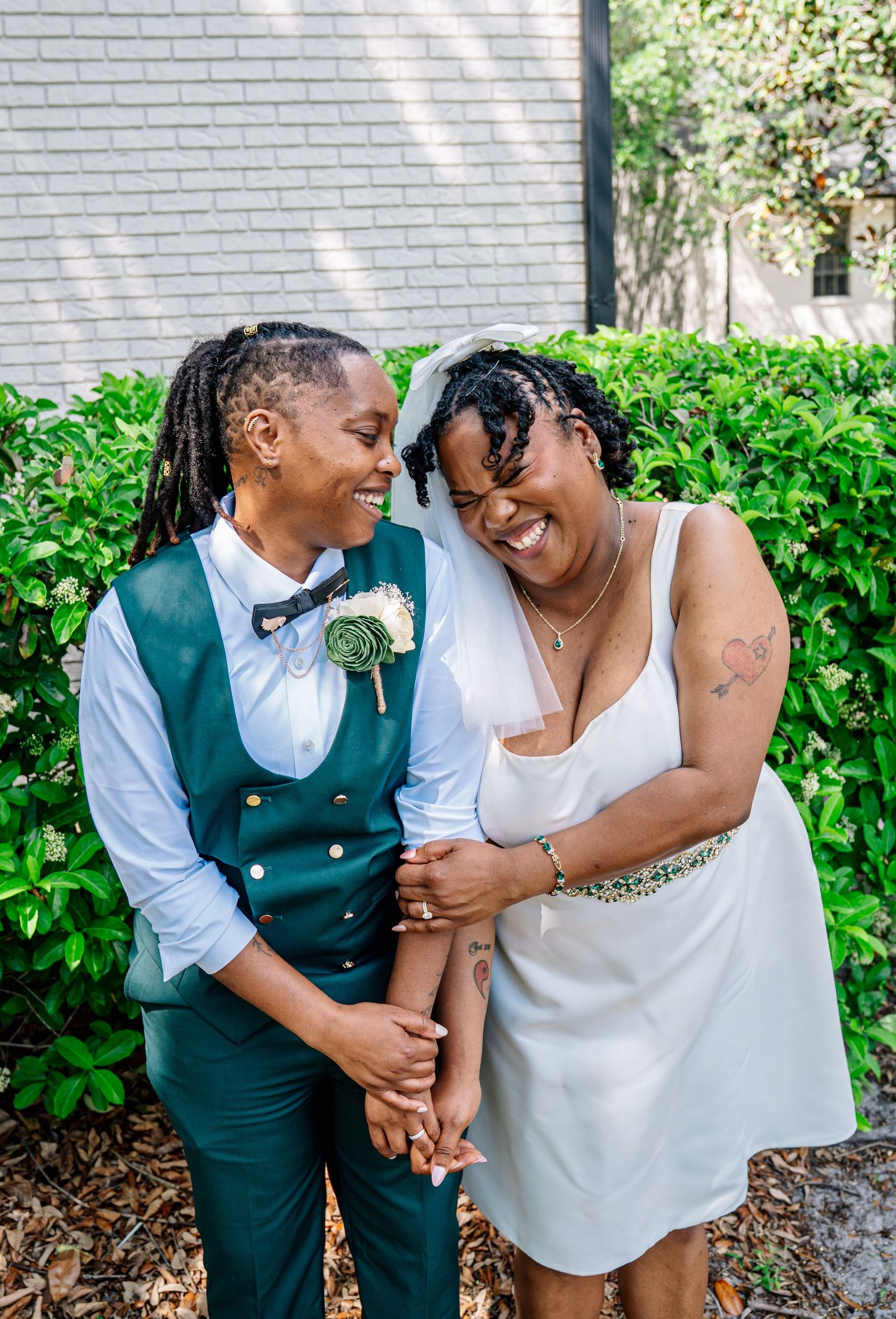 Two brides laugh joyfully outdoors, one in a white dress and one in a green vest with boutonniere, on their wedding day.