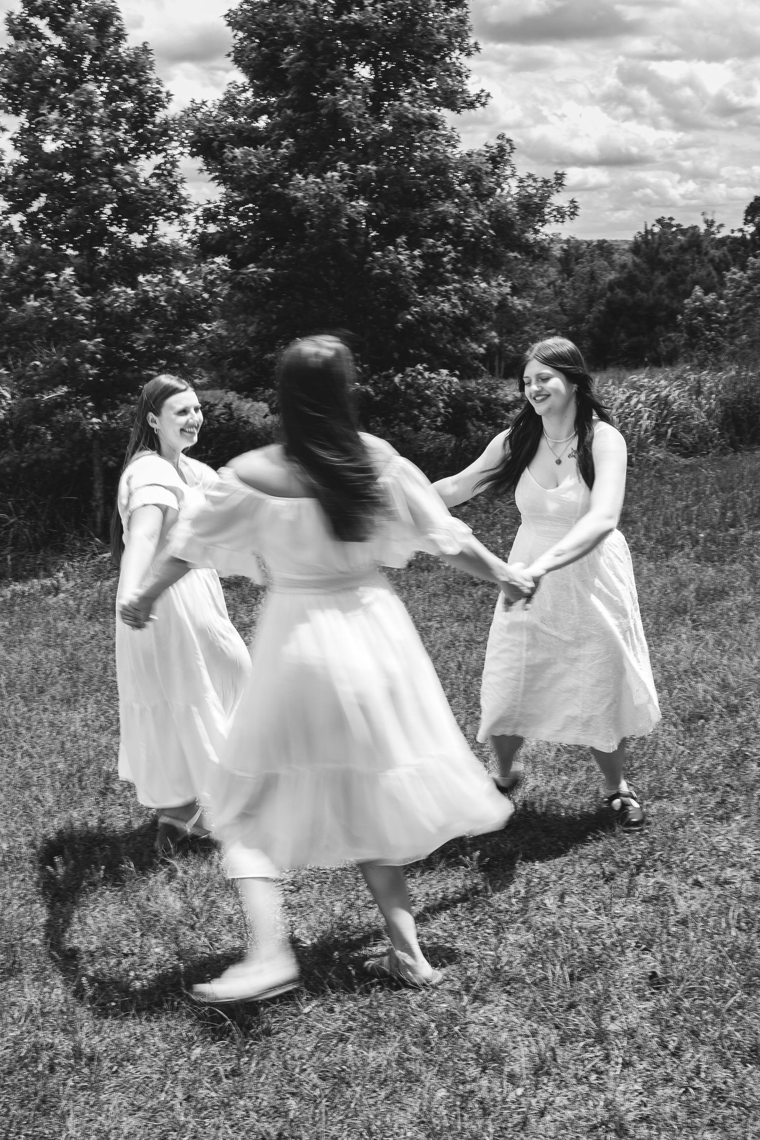 Three women in white dresses hold hands dancing in a circle outdoors in a black and white photo.