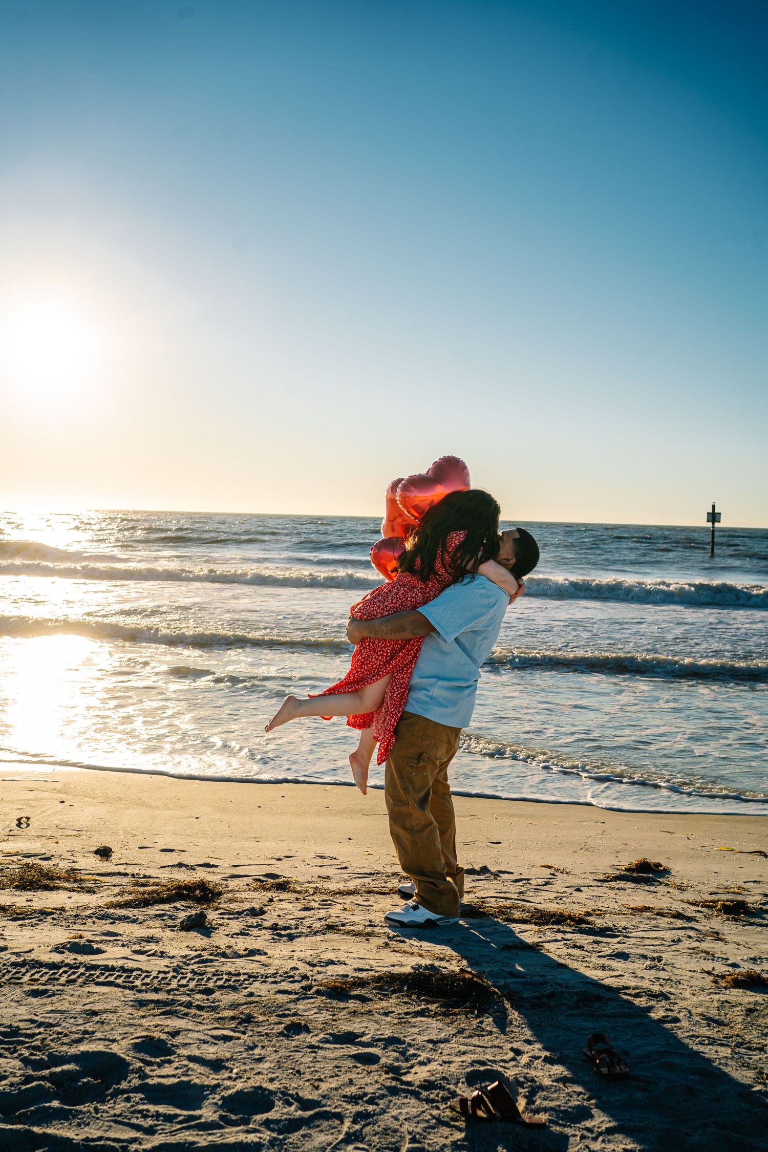 Romantic couple embracing on a sunny beach at sunset, woman in red dress held by man near ocean waves.