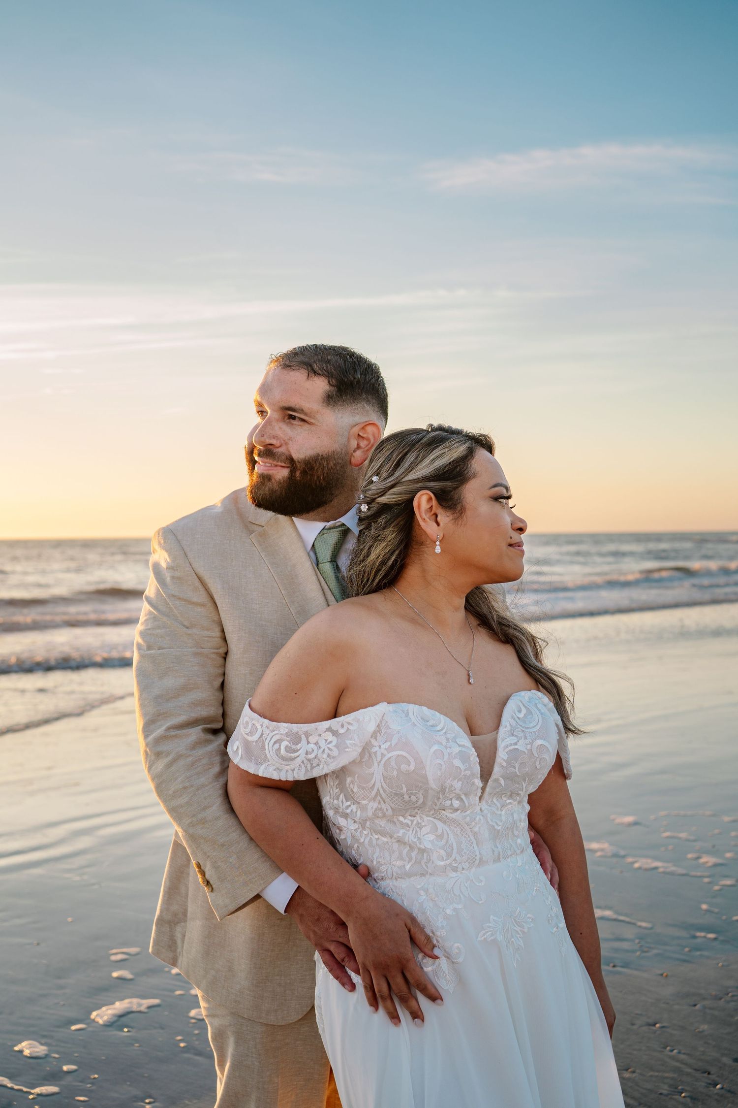 A couple in wedding attire embraces on a beach at sunset with waves and sand visible in the background.