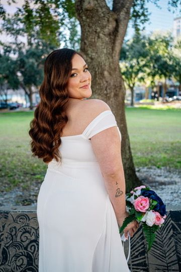 A person in a white wedding dress poses outdoors against an ornate bench with pink roses in a garden setting.