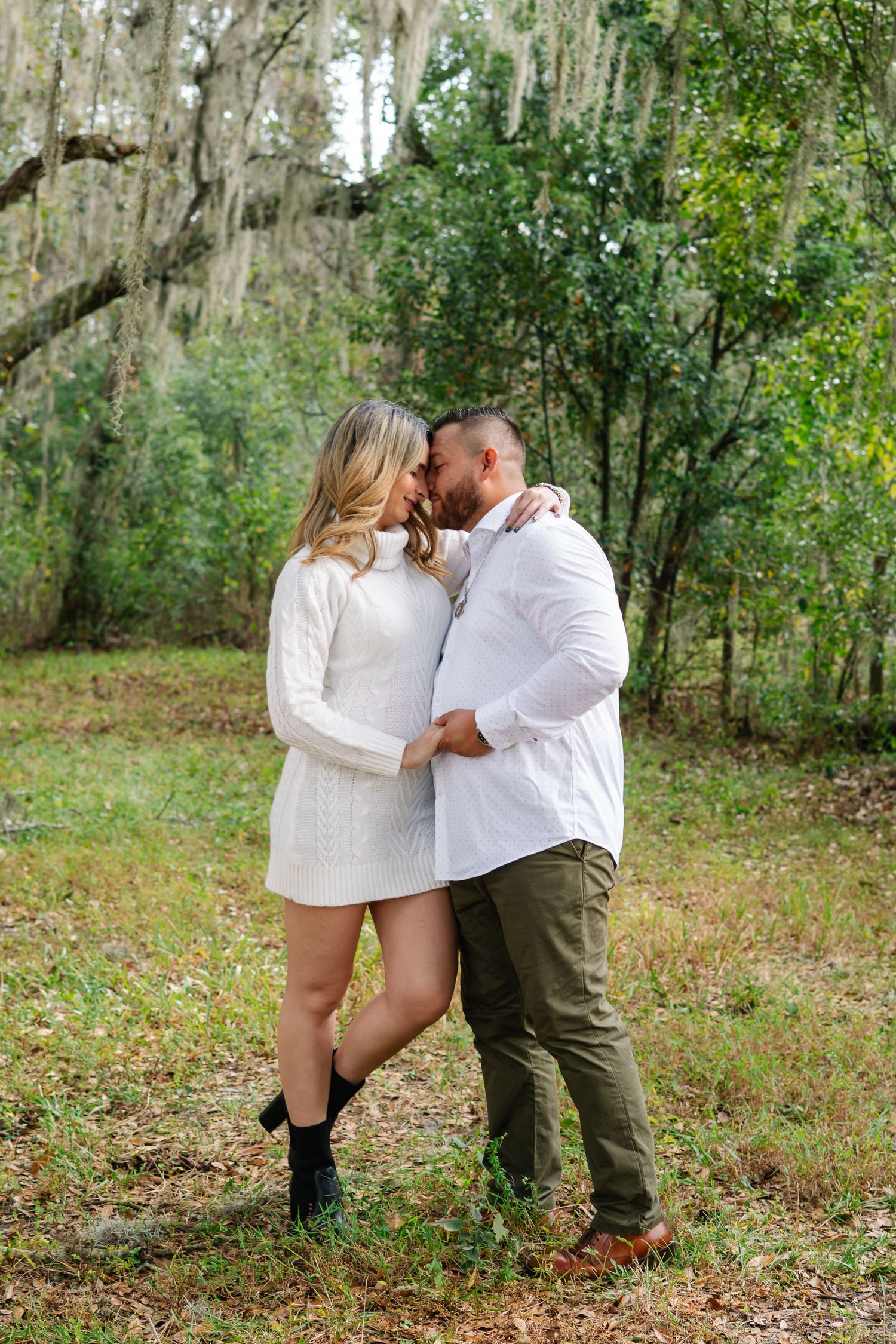 Romantic couple in white outfits share an intimate moment in a mossy wooded setting.
