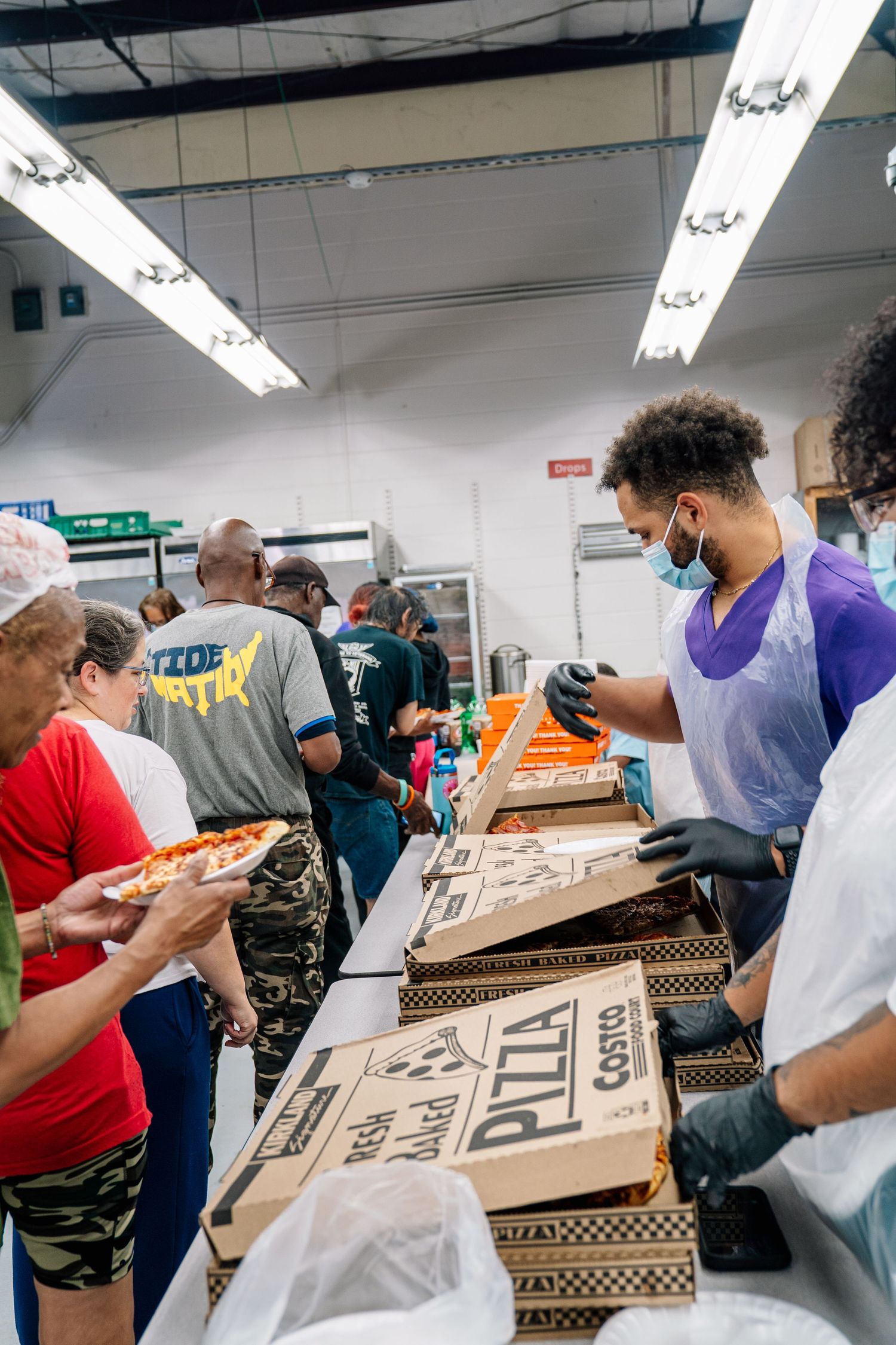 Volunteers and recipients gather around pizza boxes at a busy indoor community food distribution event.