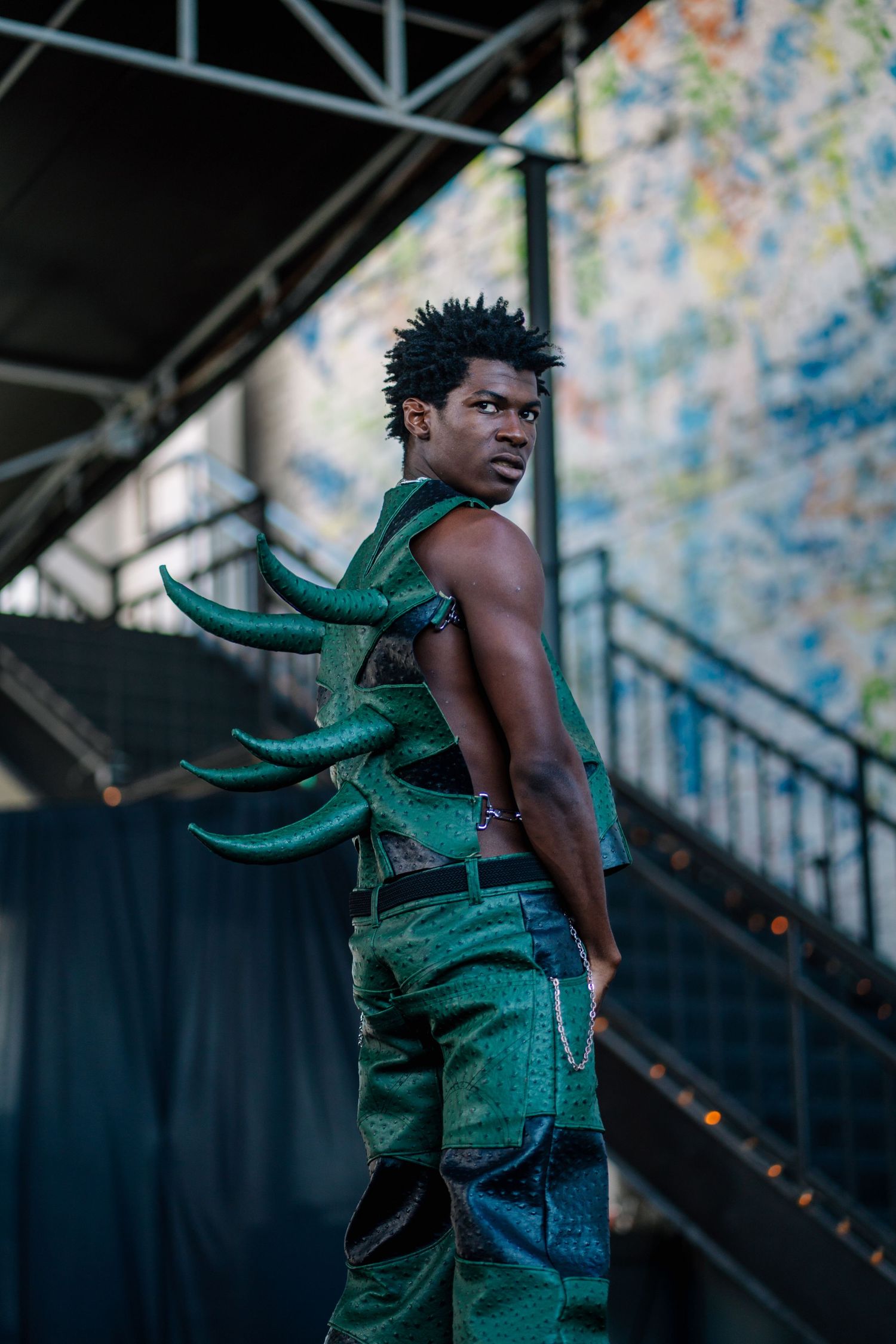 Young man in green reptile outfit with spikes poses on outdoor stage stairs at music festival.