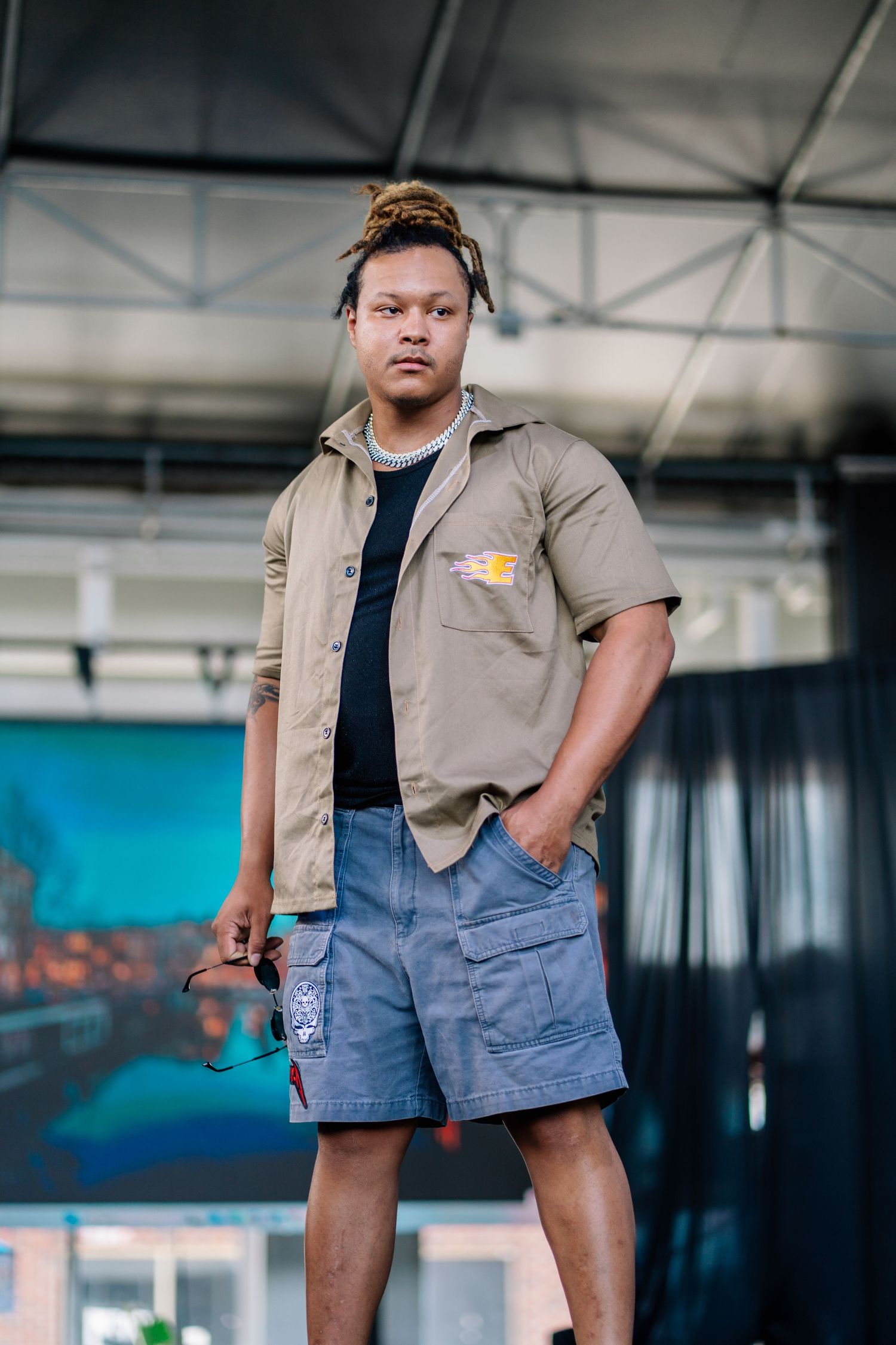 Young man with dreadlocks wearing khaki shirt and blue cargo shorts stands confidently outdoors.