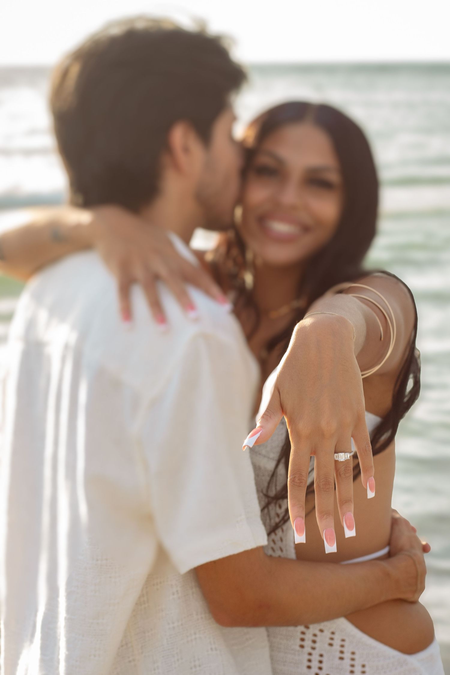 A romantic beach engagement photoshoot shows a couple embracing while displaying a ring at sunset.