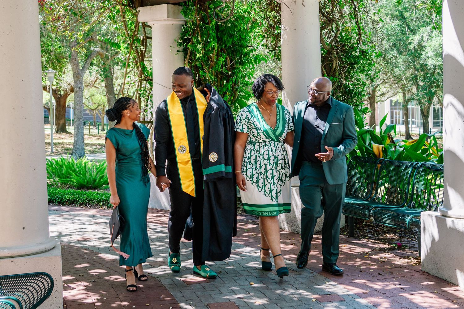 Graduate in cap and gown walks with family on college campus during graduation celebration outdoors.