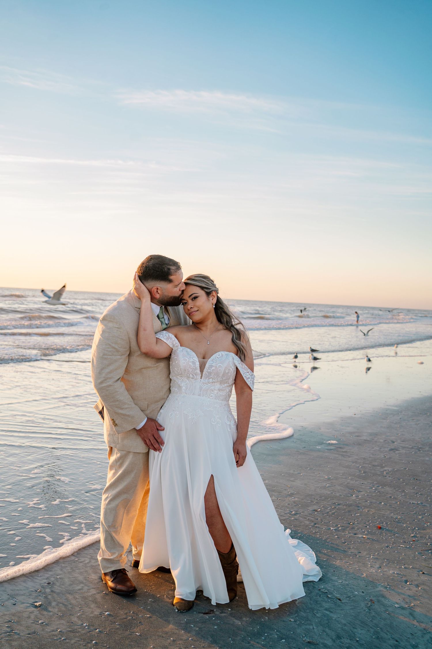 Bride and groom share a romantic moment on a beach at sunset, waves gently lapping at their feet.