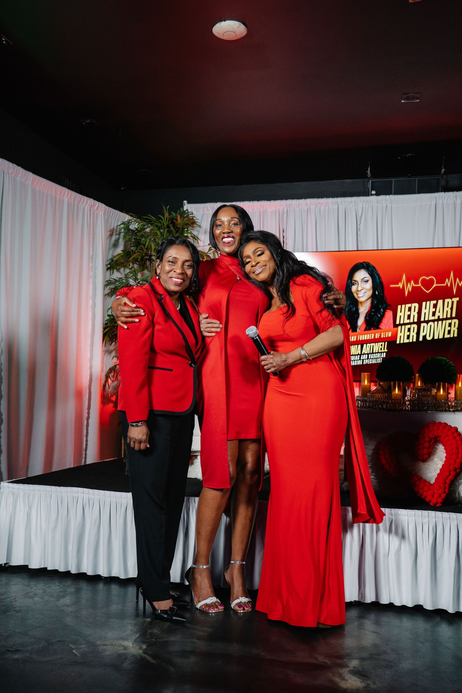 Three women in red outfits pose together at a Her Heart Her Power event, smiling on a decorated stage.
