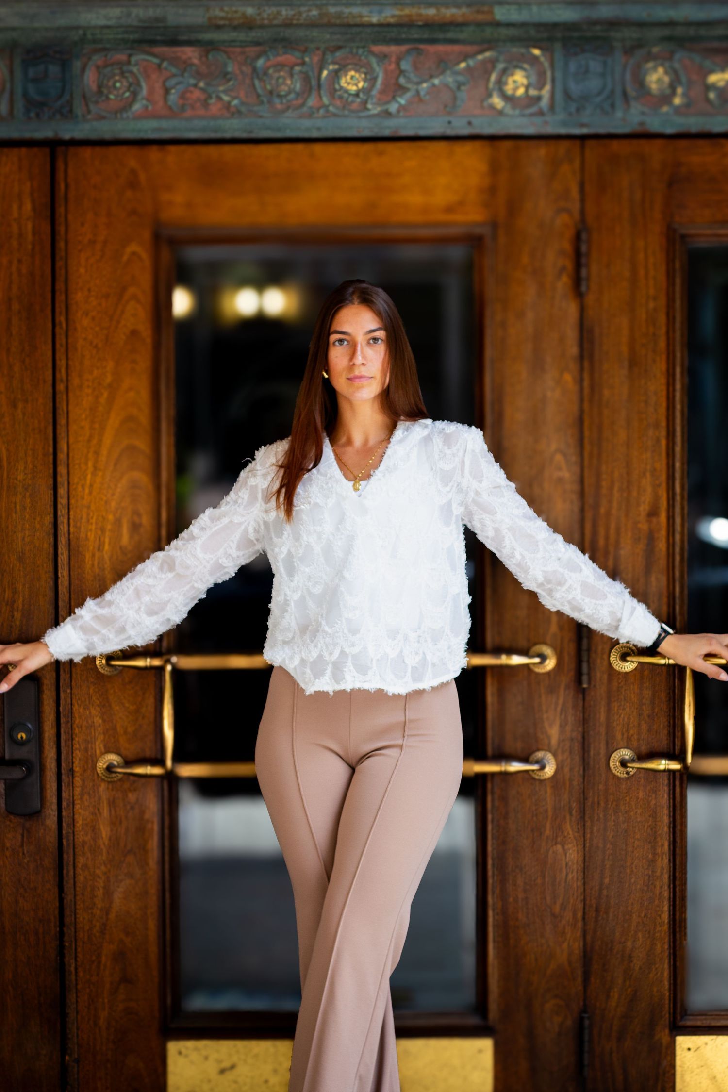 A person in a white ruffled blouse and beige pants poses against dark wood paneling in an elegant interior.
