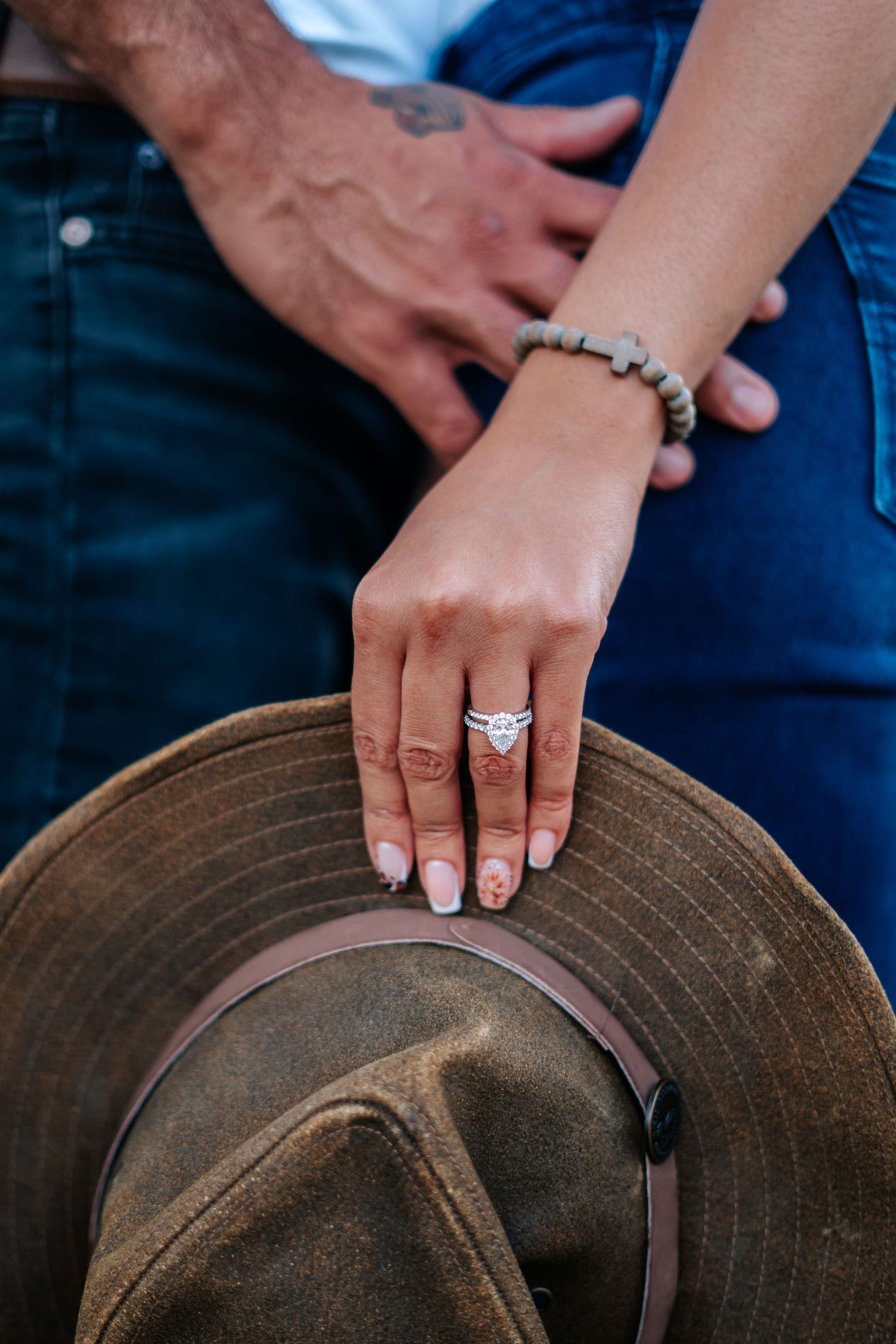 Close-up of a woman's hand wearing a diamond engagement ring resting on a brown hat, with a man's tattooed hand nearby.
