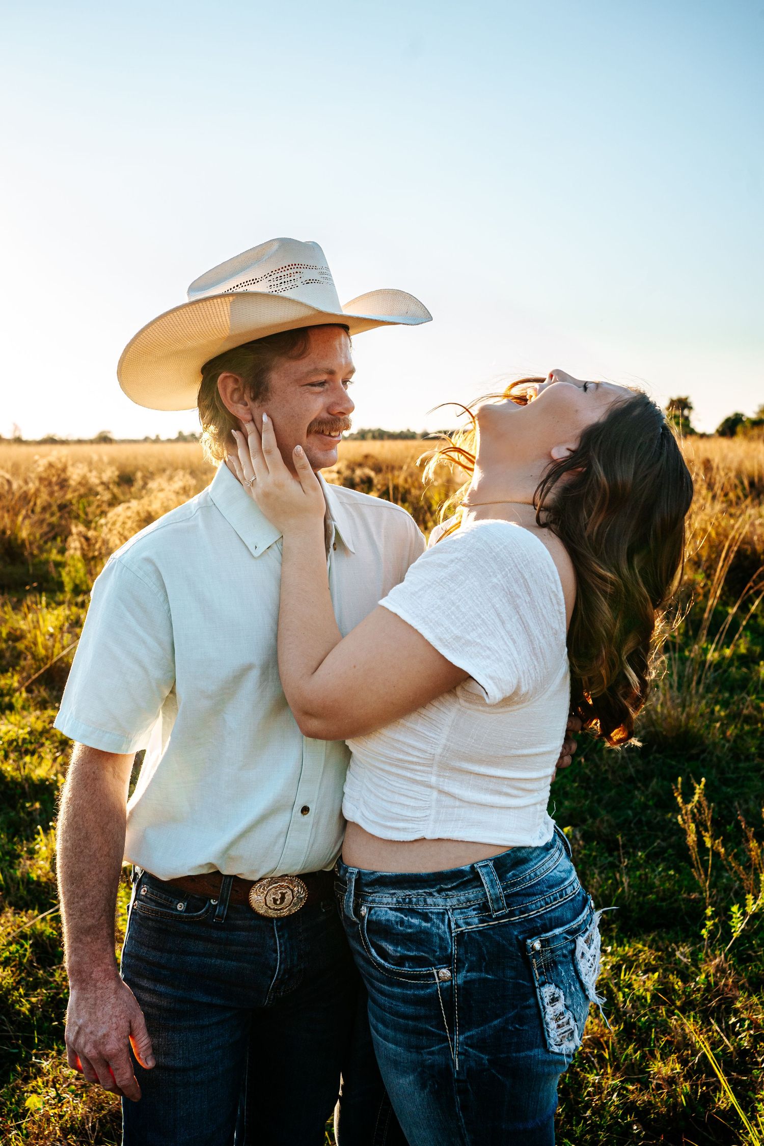 Laughing couple in golden field at sunset, woman touching mans face as he wears a white cowboy hat.