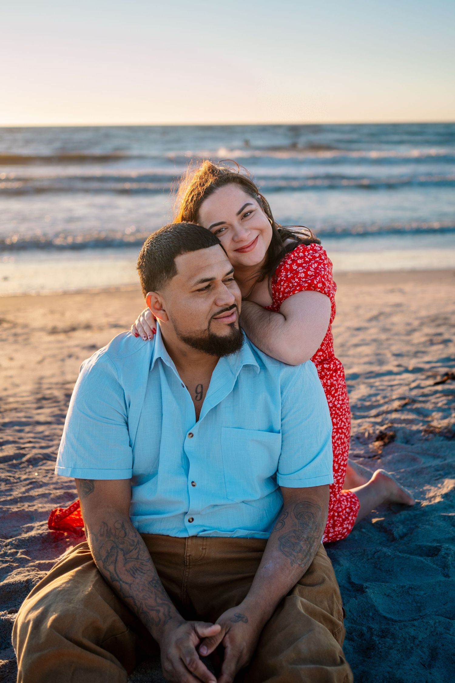 Couple sitting on sandy beach at sunset, woman in red dress leaning on man in blue shirt, ocean waves behind them.