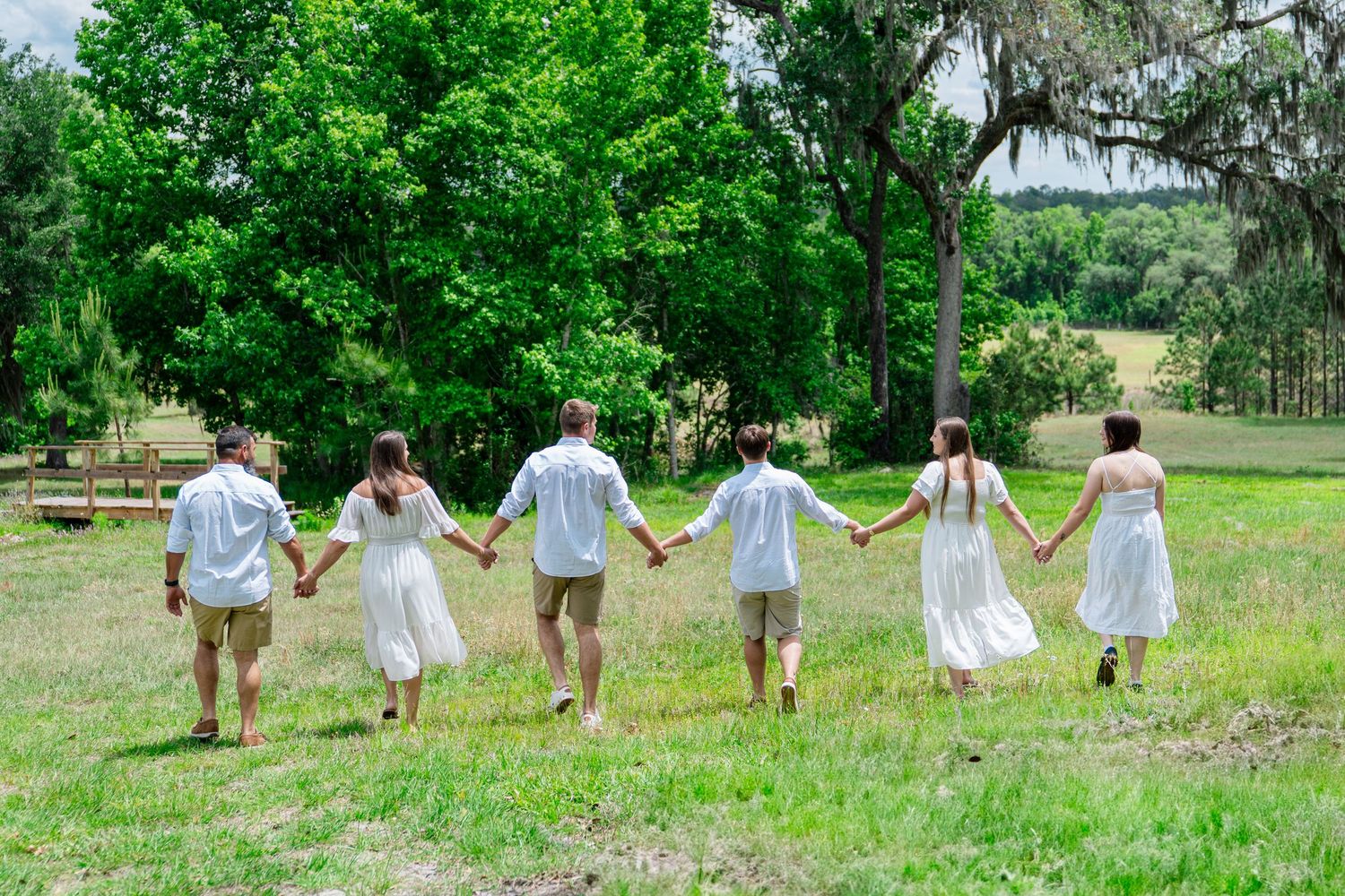 Six people in white and khaki outfits hold hands walking through a lush green field surrounded by trees.