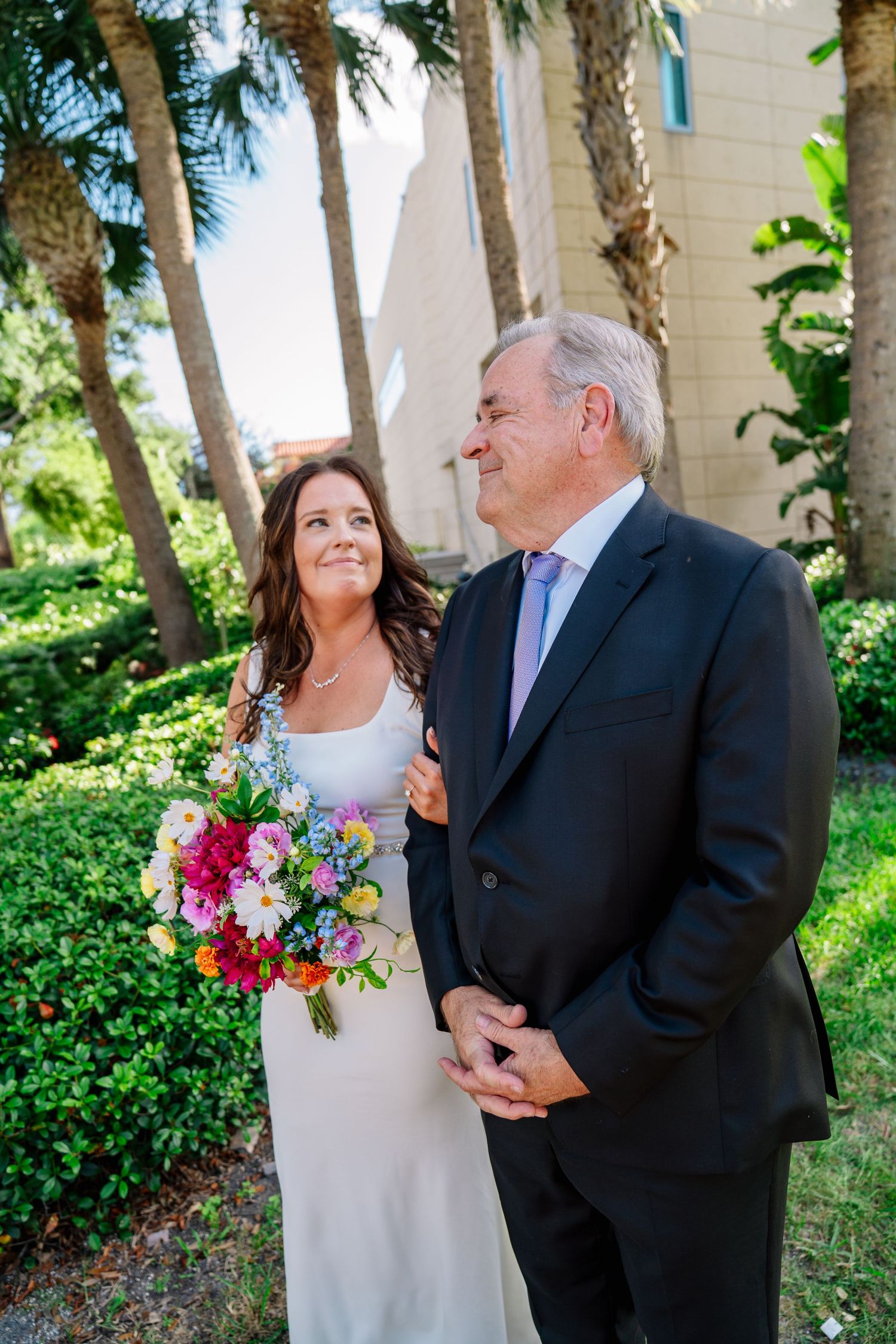 Bride in white gown holding colorful bouquet stands with older man in dark suit outdoors near palm trees.