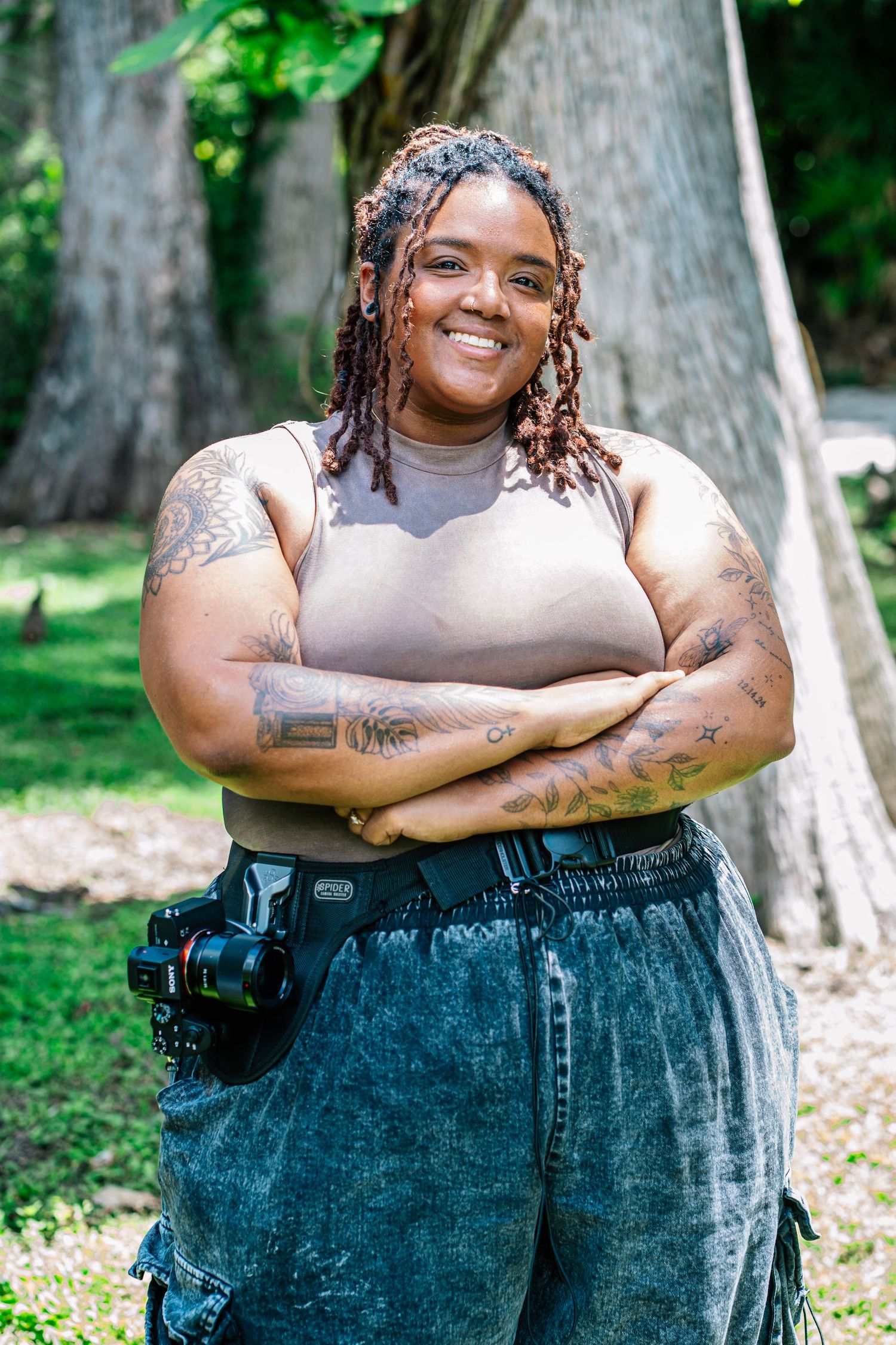 Smiling woman with tattoos and locs, arms crossed, wearing a crop top and jeans with a camera bag outdoors.