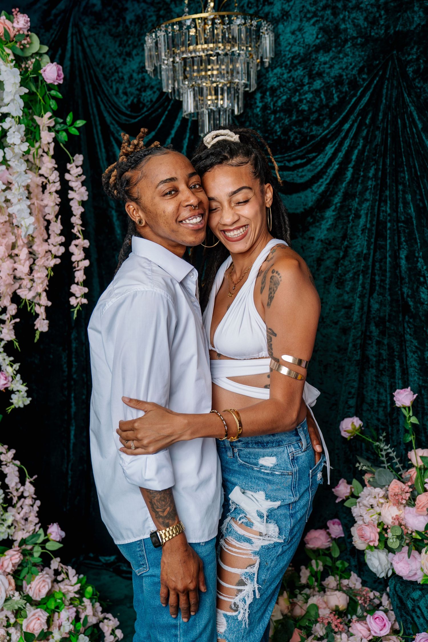 A smiling couple embraces in front of a teal velvet backdrop with pink flowers and a crystal chandelier.