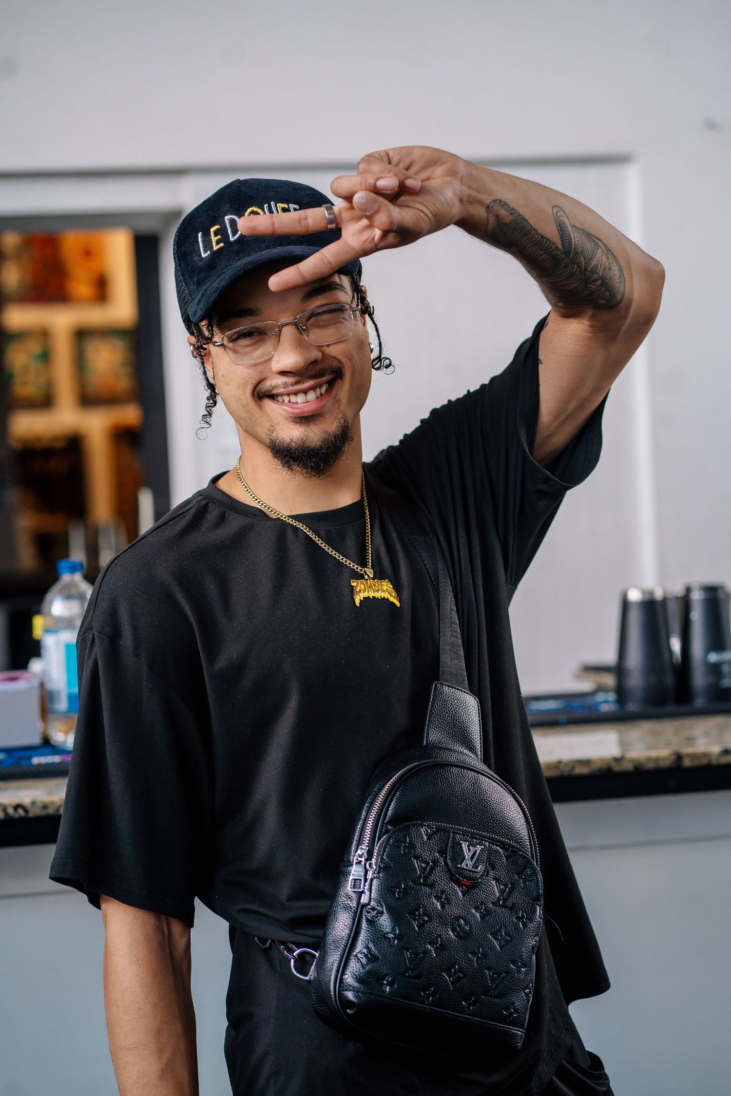Smiling young man wearing black outfit, Louis Vuitton sling bag, gold chain, cap, and glasses making peace sign.