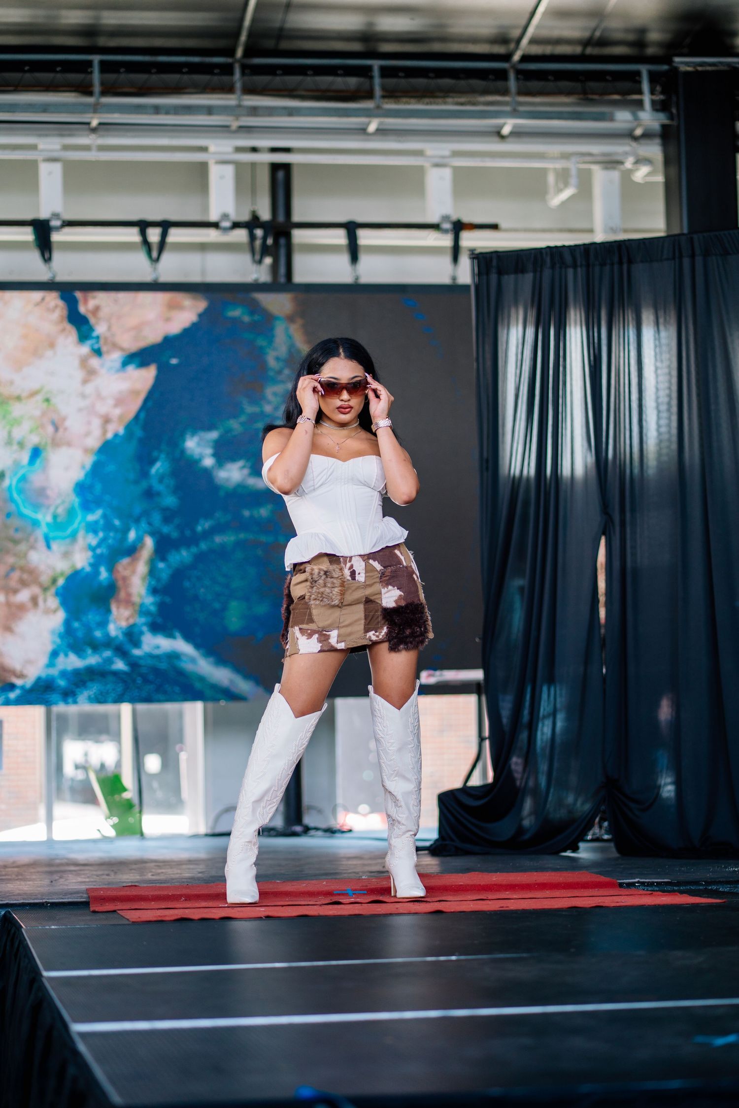 A woman in white top, camo skirt, and thigh-high boots poses on a runway stage with a world map backdrop.