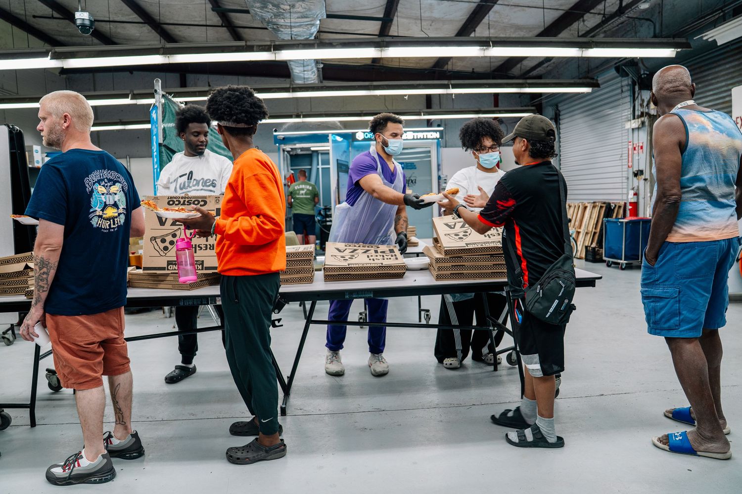 People wearing masks gather around a table in a warehouse, engaged in a community food or supply distribution event.