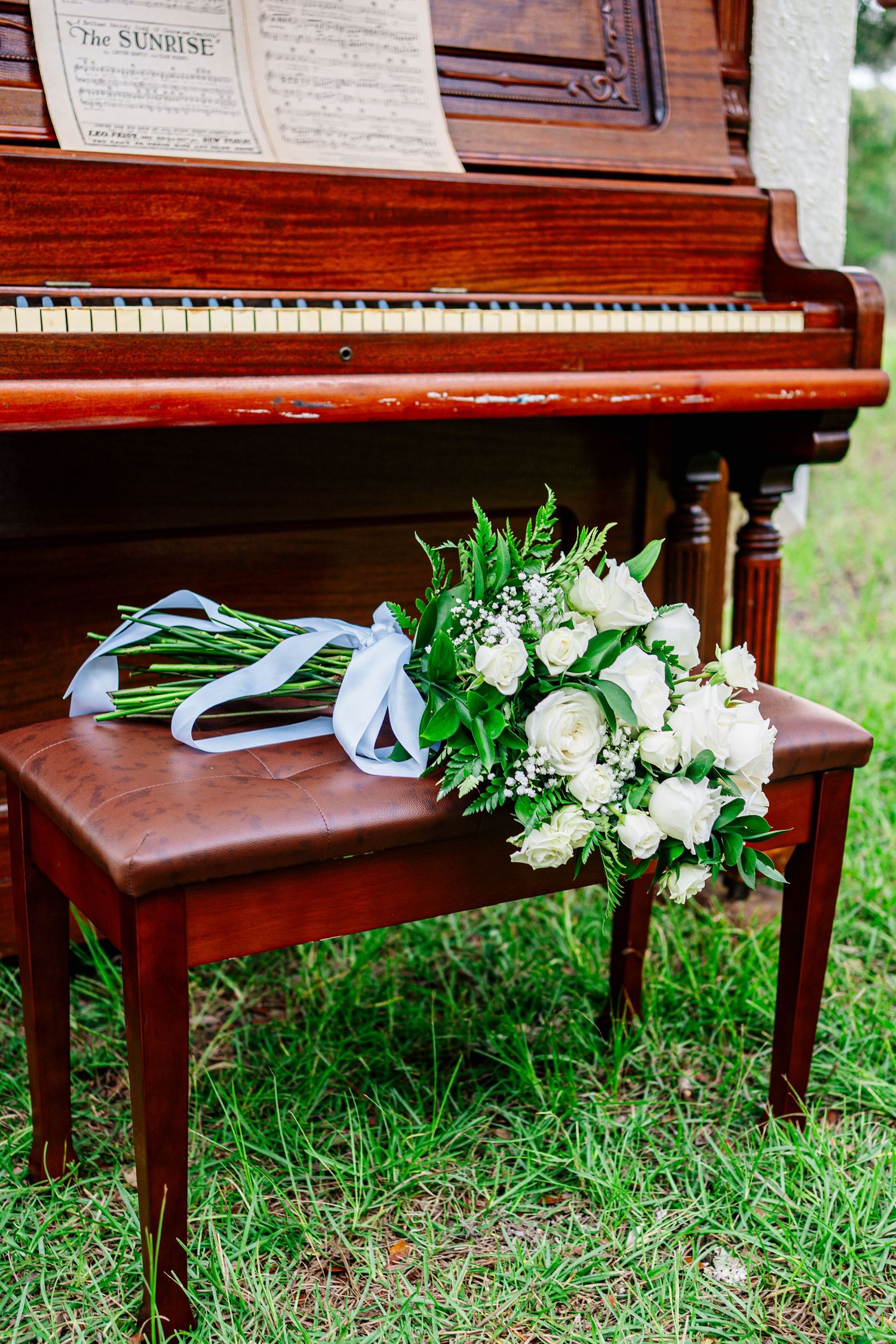 White rose bridal bouquet with blue ribbon resting on a wooden piano bench beside a vintage upright piano outdoors.
