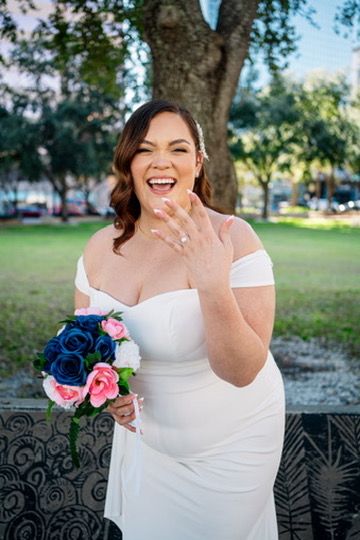 Joyful bride in white off-shoulder dress laughs while holding blue and pink bouquet in outdoor garden setting.