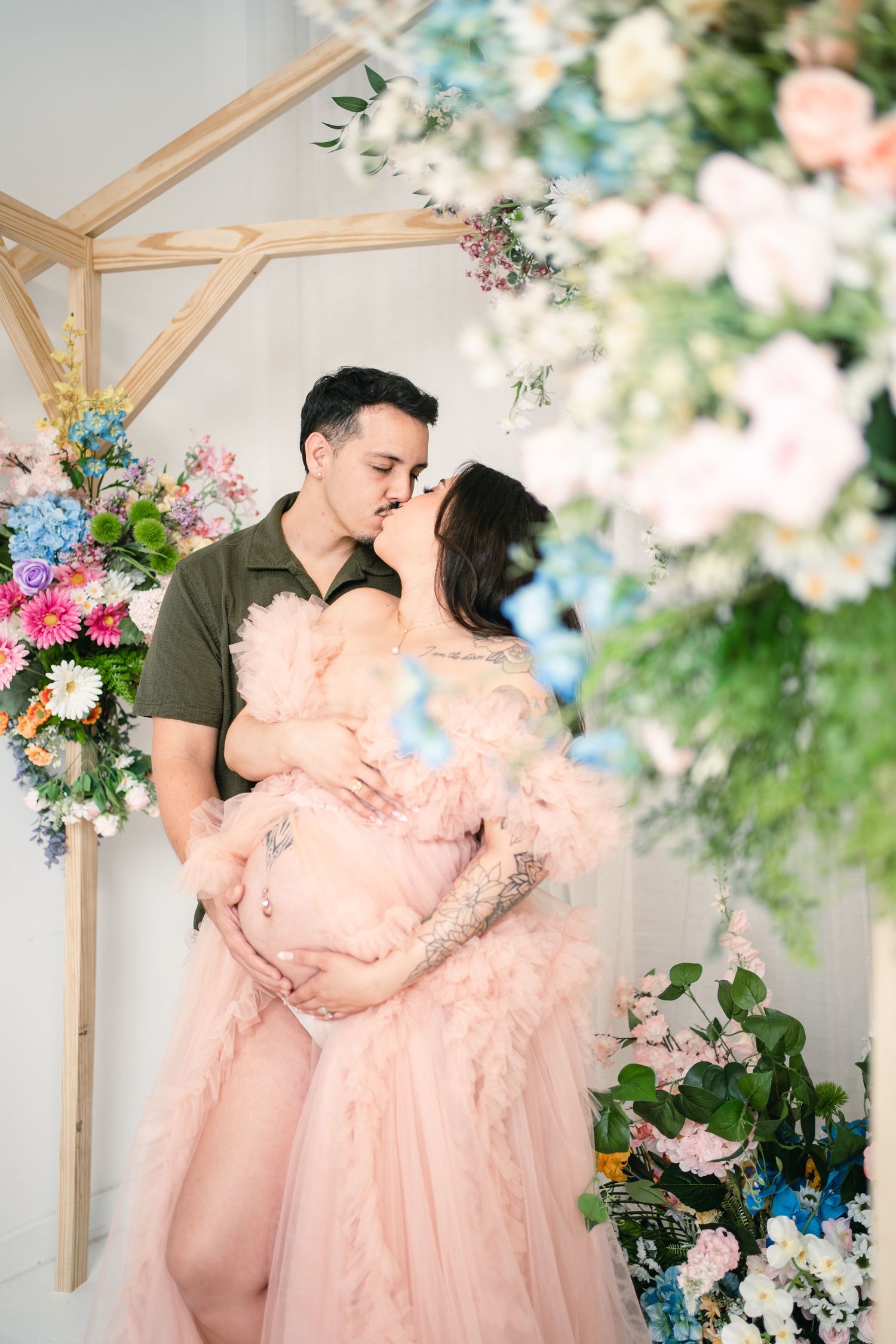 Couple kissing during maternity photoshoot, pregnant woman in pink tulle gown, floral arch backdrop.