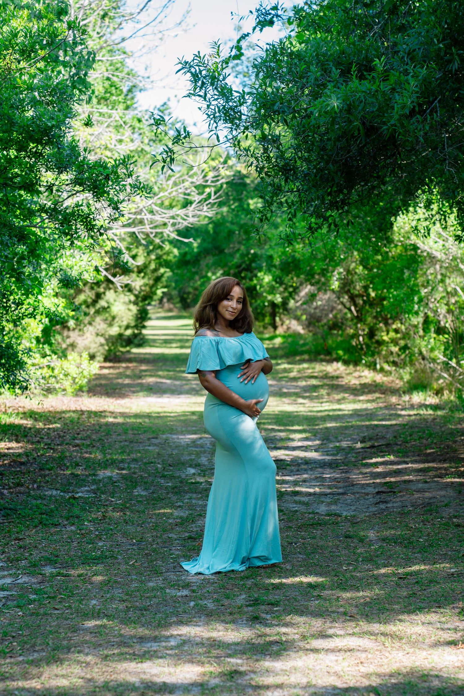 Pregnant woman in teal off-shoulder gown posing on a tree-lined path during an outdoor maternity photoshoot.