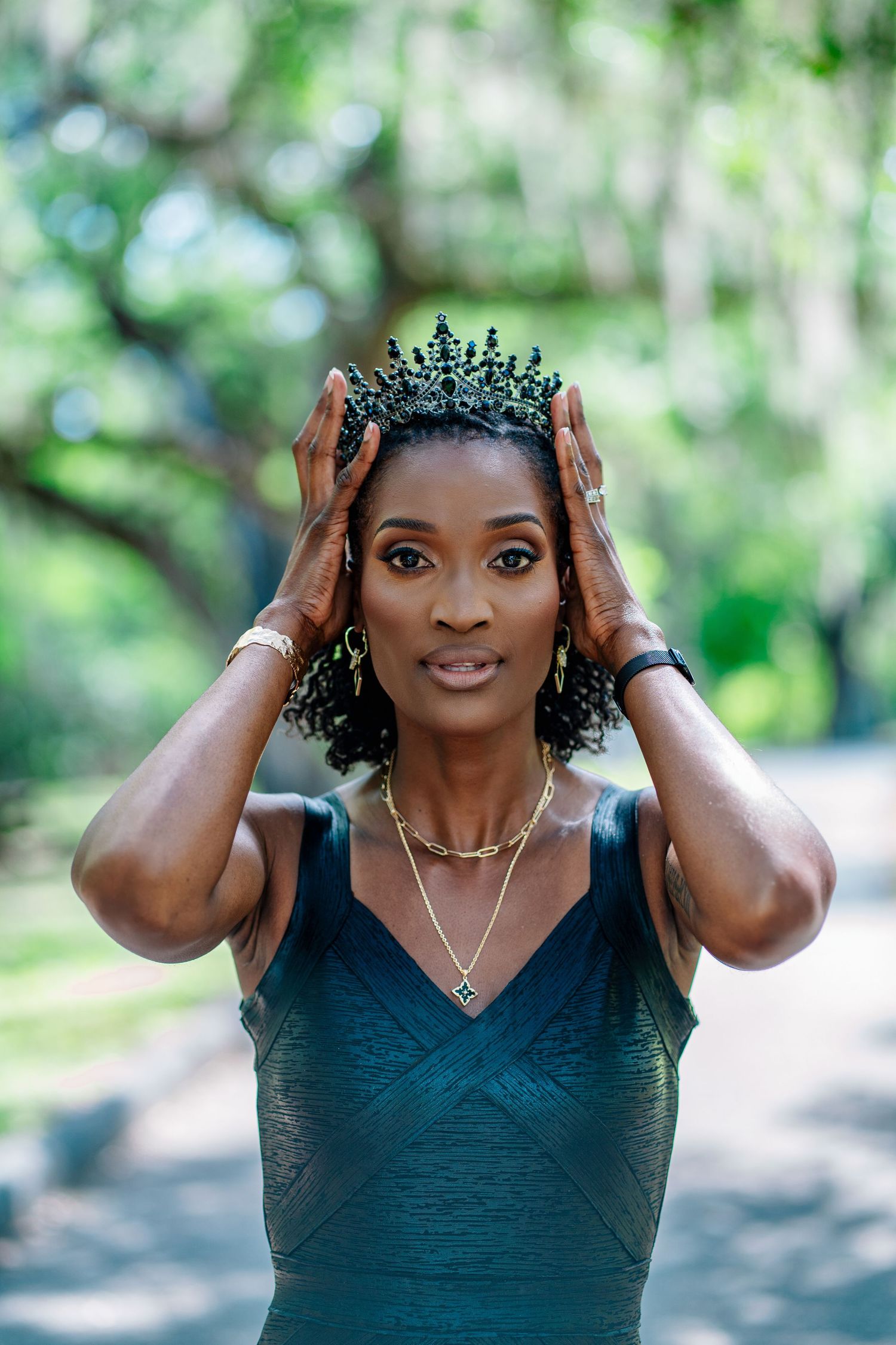 A Black woman in a teal dress adjusts a dark crown on her head outdoors in a lush green park setting.