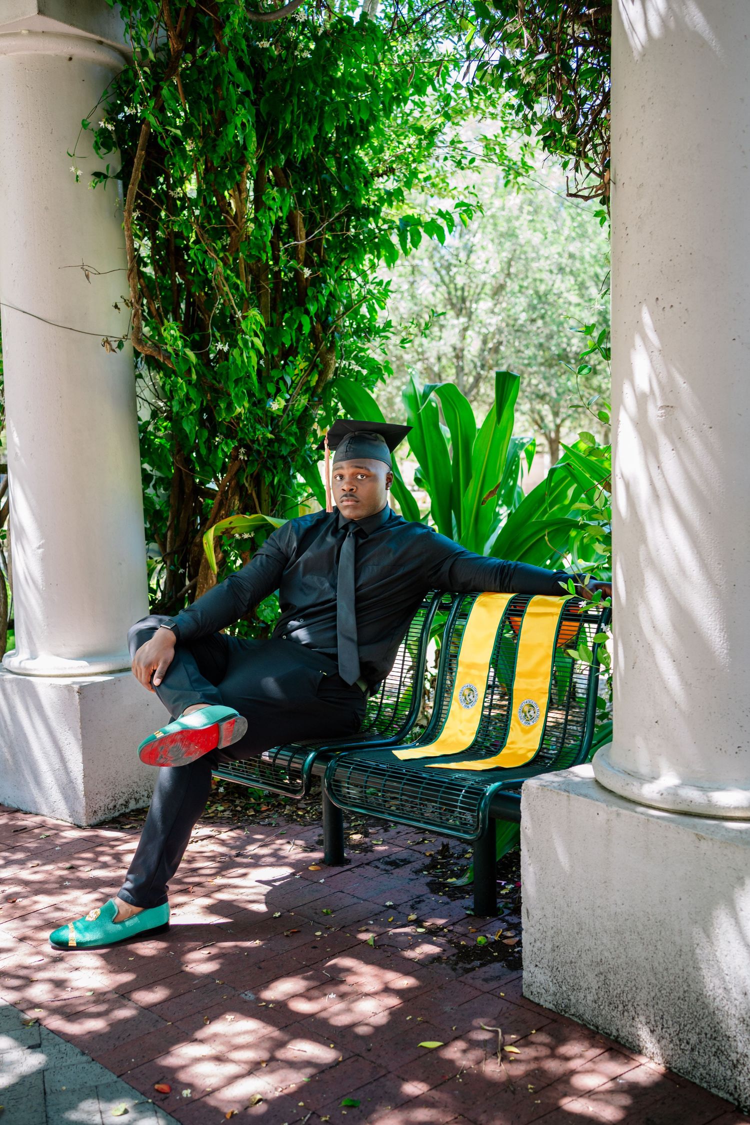 Graduate in cap and gown sits on green bench surrounded by lush greenery, wearing teal shoes with red soles.