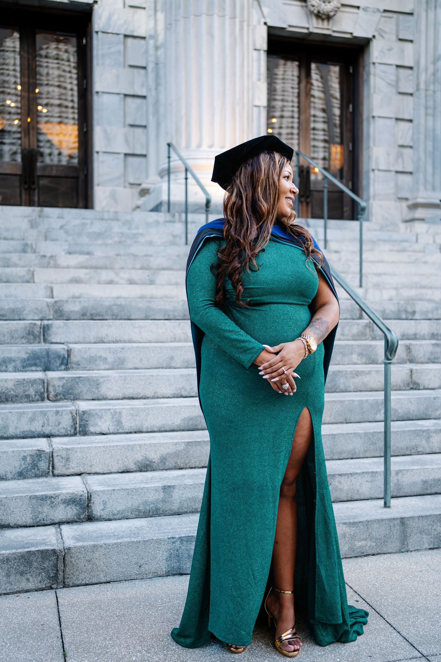 Woman in green gown and graduation cap poses on stone steps, celebrating academic achievement.