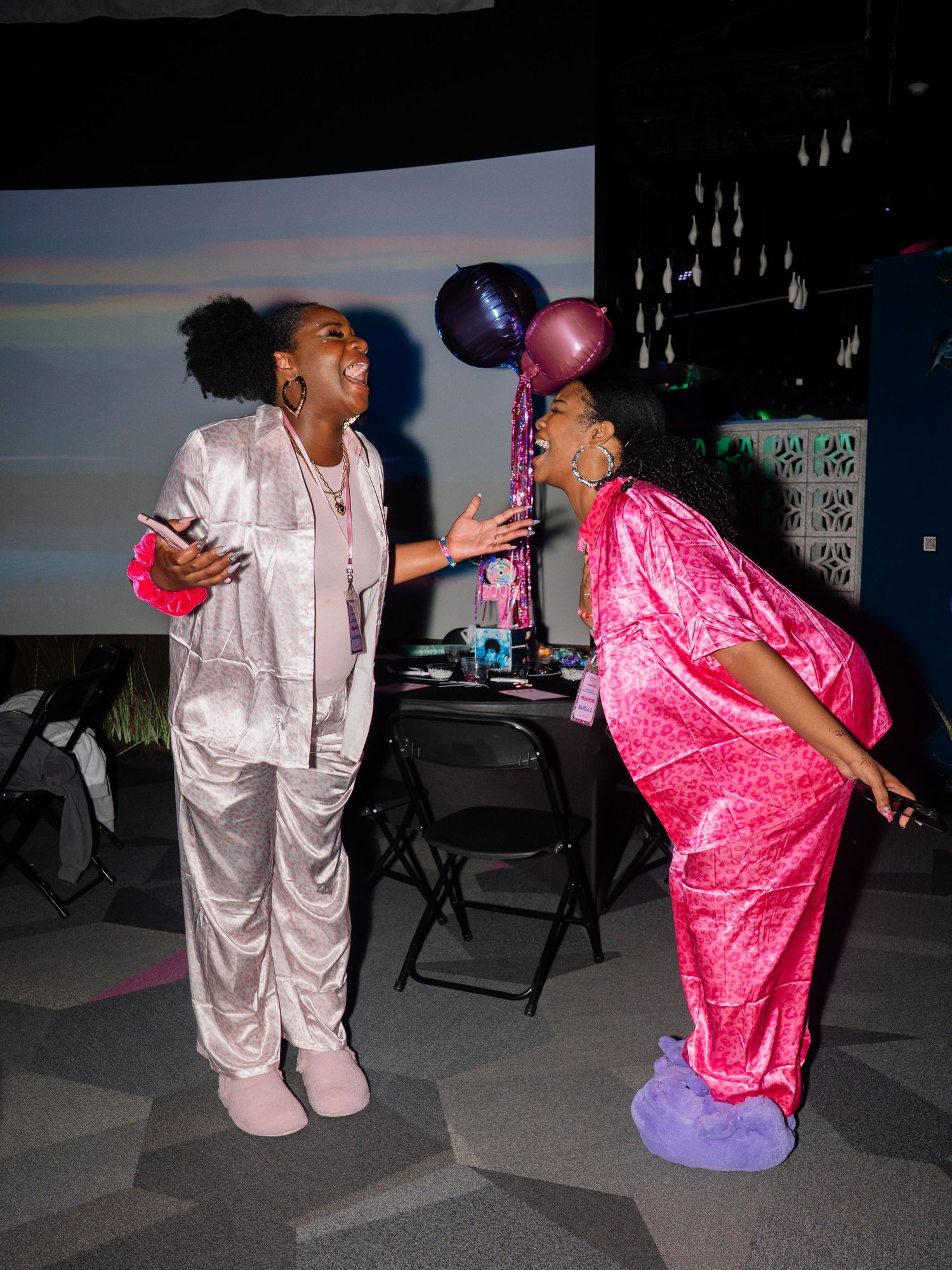 Two women in satin outfits celebrate at a party, one in pink holding balloons, laughing together indoors.