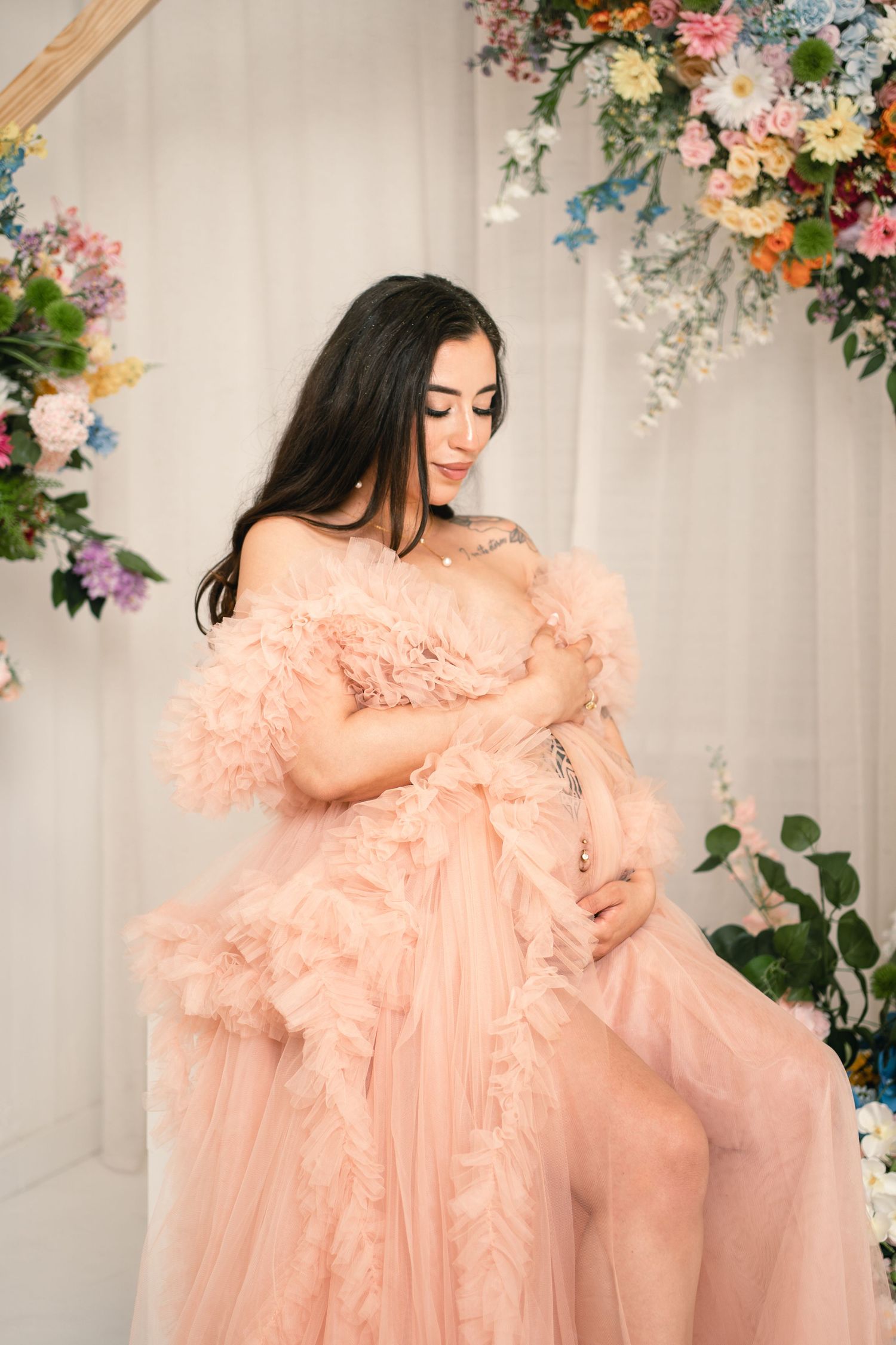 Pregnant woman in a blush pink ruffled off-shoulder gown poses gracefully among colorful floral decorations.