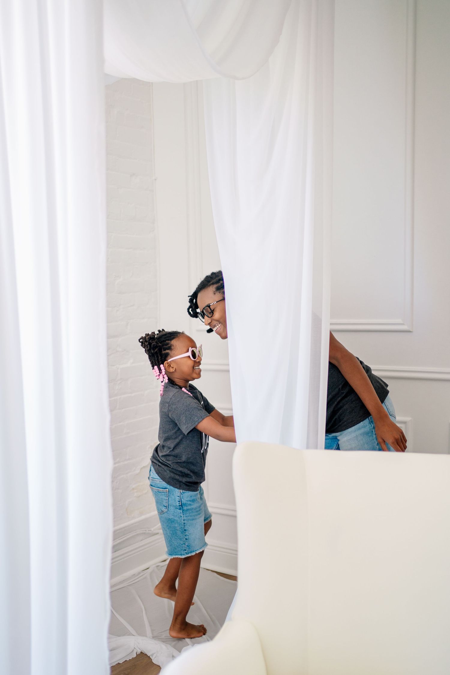 A young girl in sunglasses plays peek-a-boo with her mother behind white curtains in a bright white room.