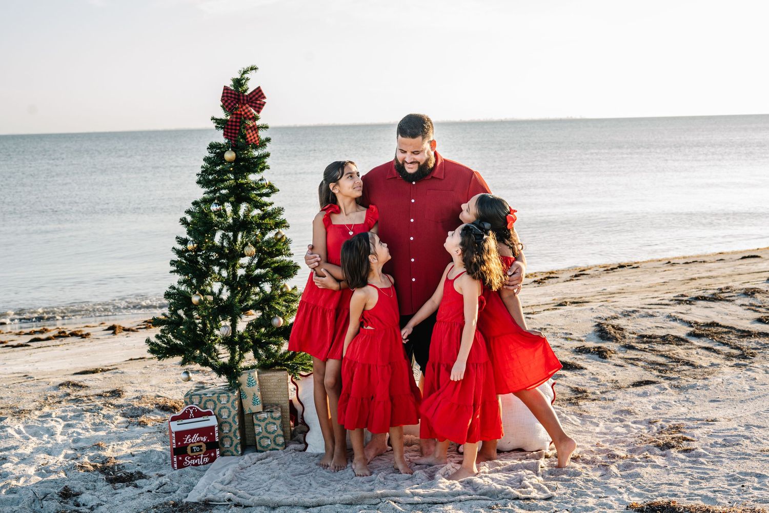 Family in red outfits posing by a decorated Christmas tree on a sandy beach during sunset.
