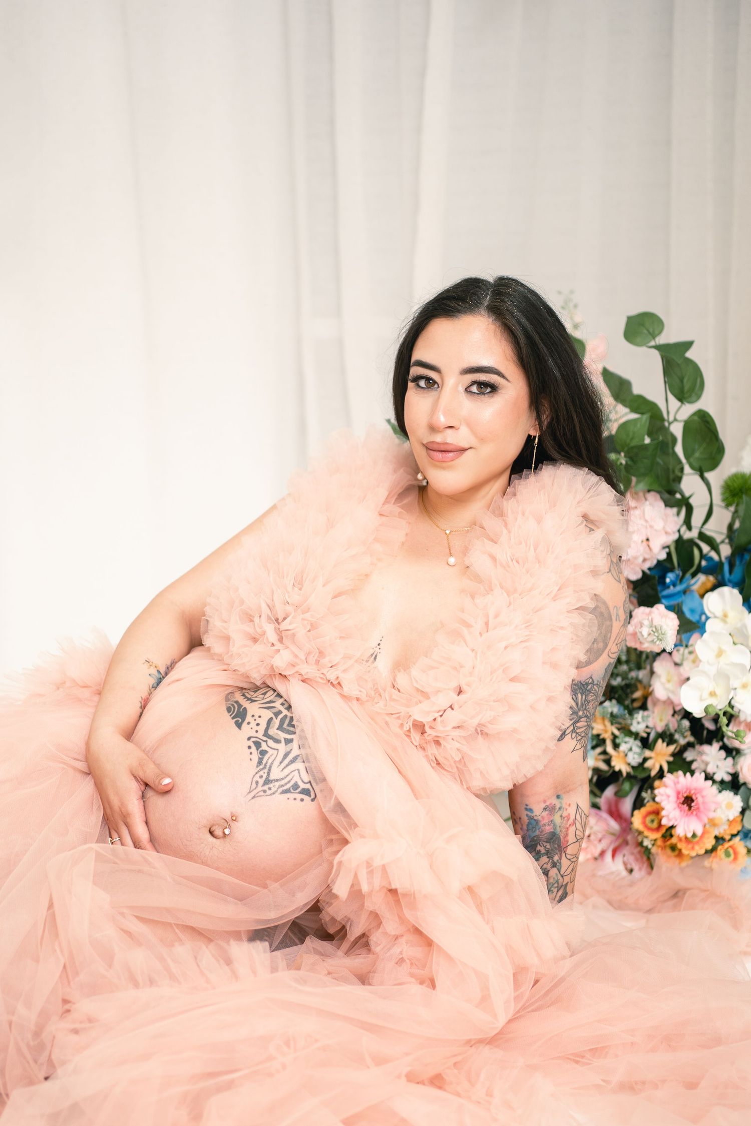 Pregnant woman in pink tulle robe posing with floral arrangement, showcasing baby bump and tattoos in a studio setting.