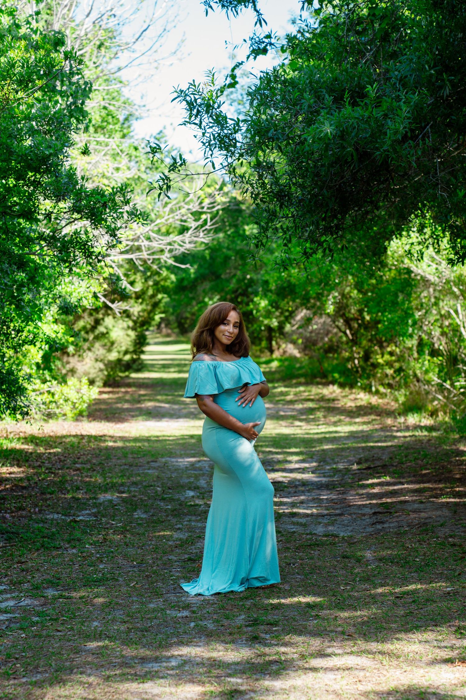 Pregnant woman in a flowing teal off-shoulder maternity gown poses on a serene tree-lined path.