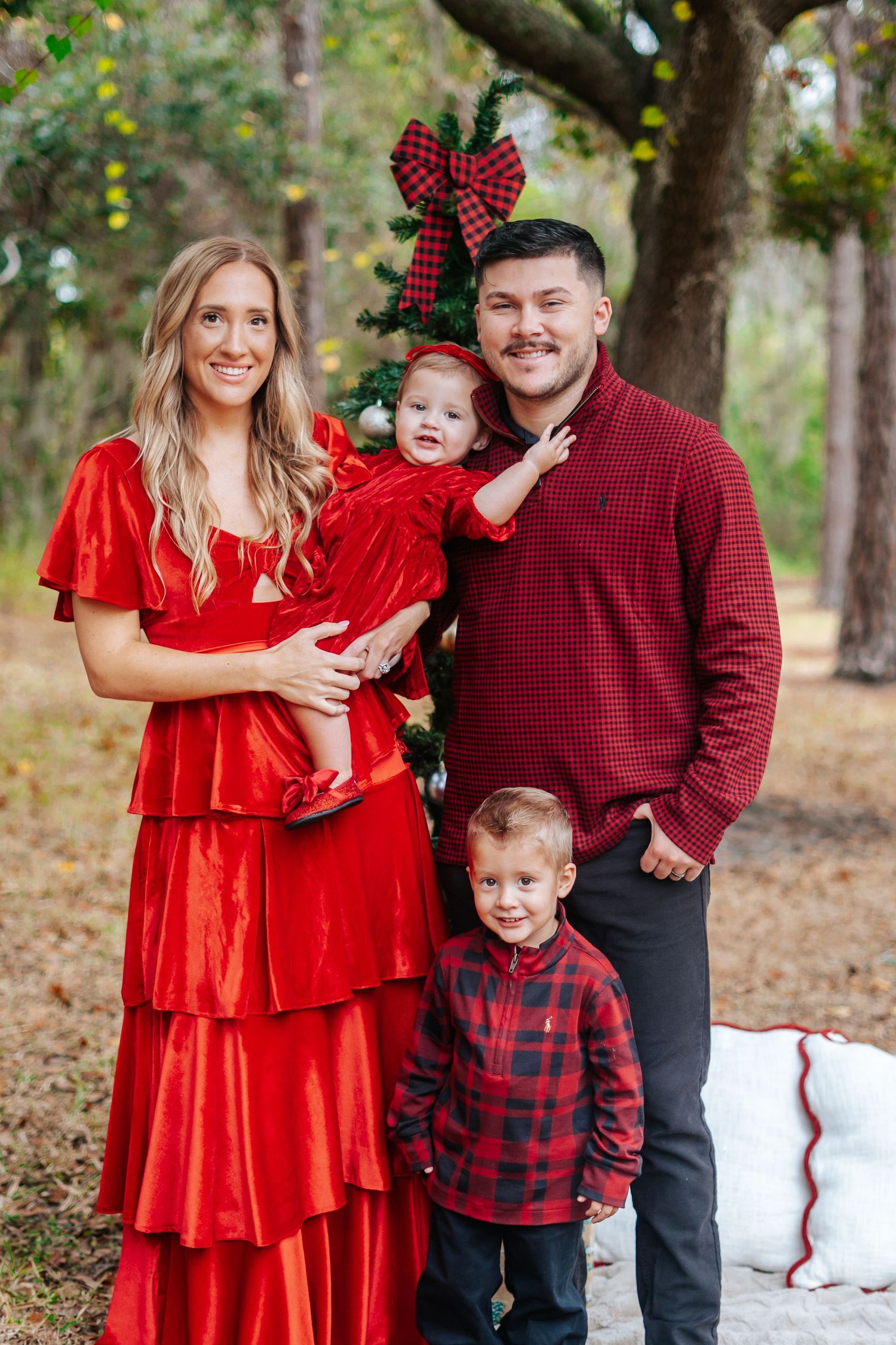 Holiday family portrait in coordinated red plaid outfits standing among trees.