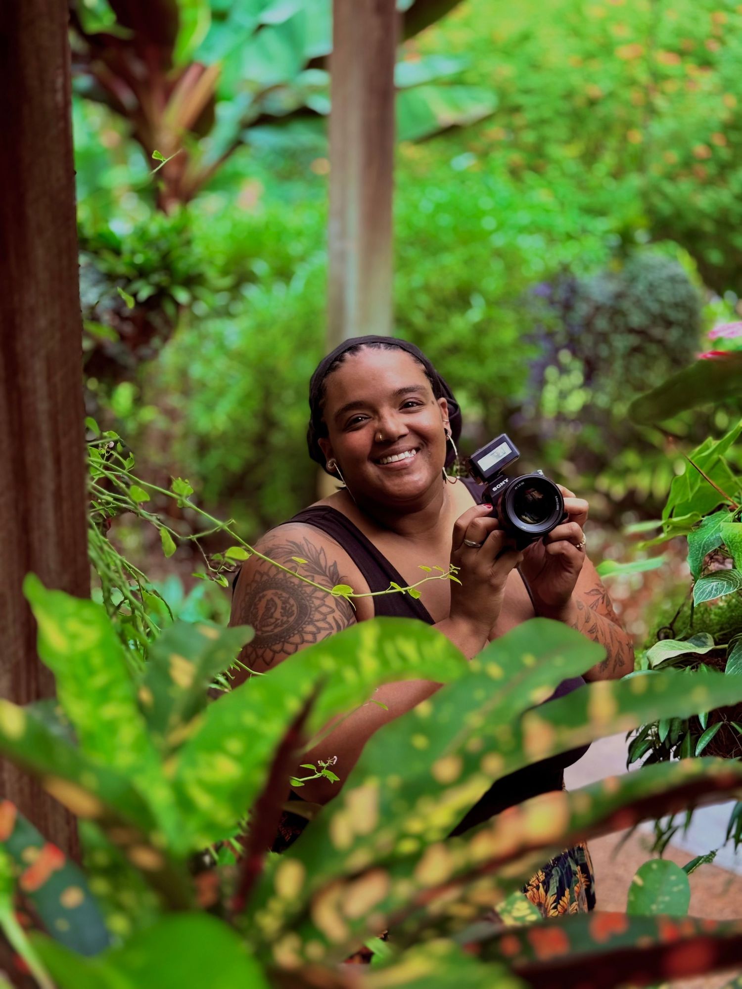 A photographer peers through lush green foliage with a camera while taking nature photos in a forest setting.