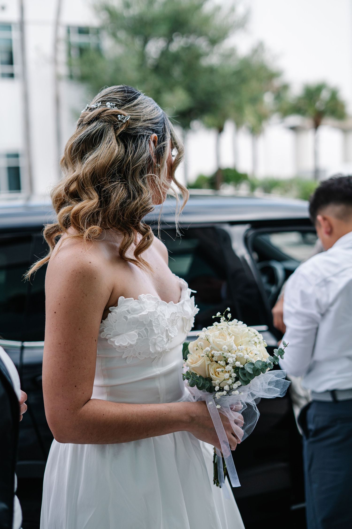 A bride in a white wedding dress holds a small bouquet of white flowers while walking outside.