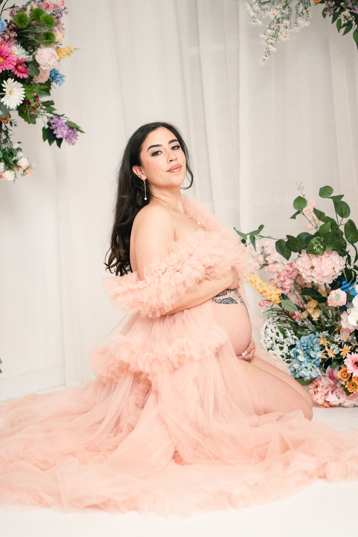 Pregnant woman in pink ruffle tulle gown poses gracefully among colorful floral arrangements in a bright studio setting.